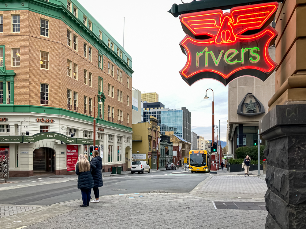 The corner of Collins Street and the Hobart Bus Mall, from just behind the Rvers store's neon sign.Two people are waiting to cross Collins Street