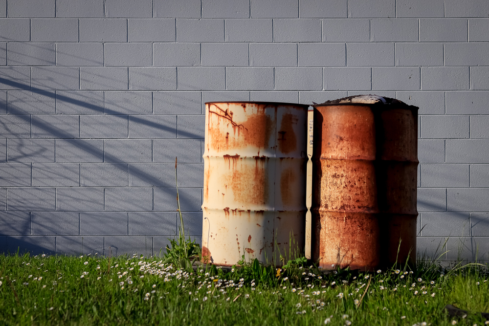 Two rusty drums standing on grass, against a grey wall
