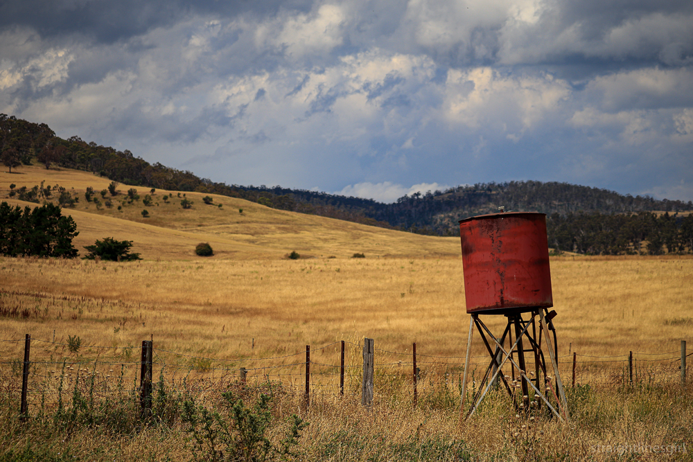 A rusty water tank in the foreground of dry hills, a fence and looming clouds