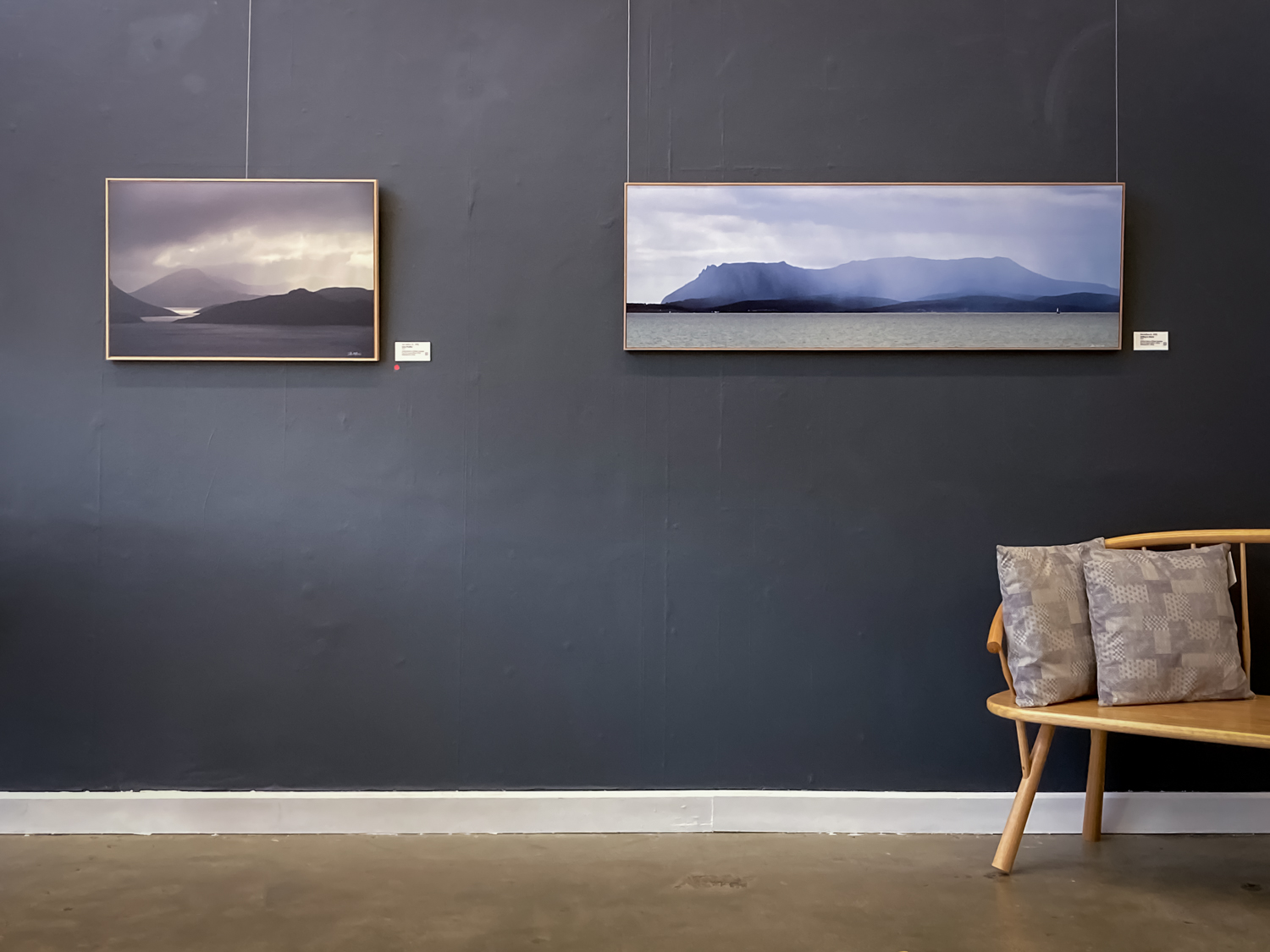 Two landscape photographs hung on a dark grey wall with a cane chair in the foreground