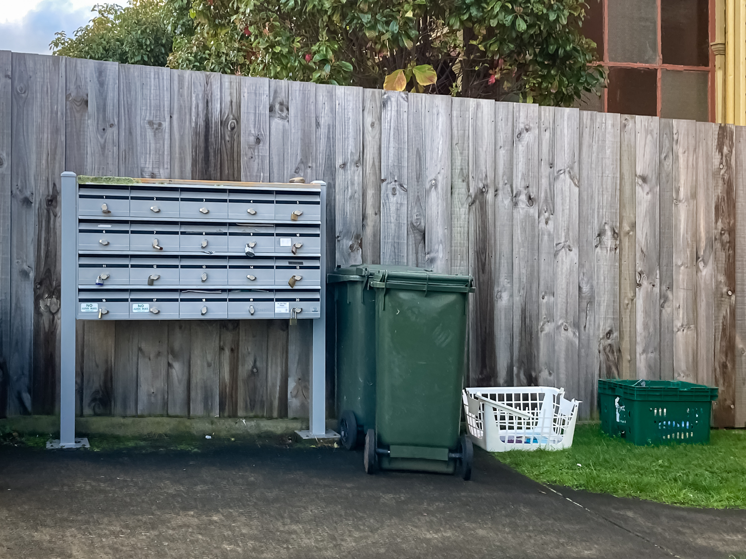 A silver multi-box letterbox, green bins and a white plastic basket in front of a fence