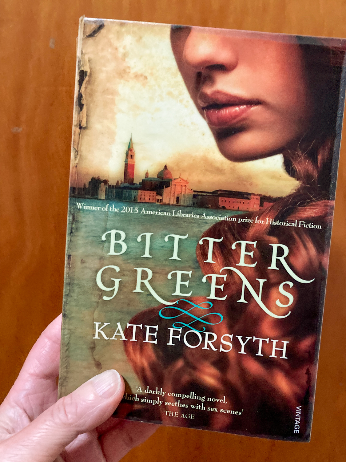 A hand holding a book title Bitter Greens by Kate Forsyth with a close up of a woman's face and red hair