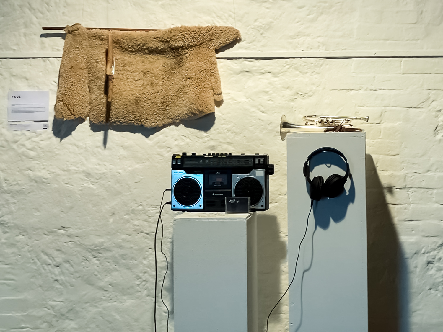 A white wall. A sheepskin with a knife hanging in front of if. Two plinths: one with a cassette player, the other with a brass instrument and some headphones