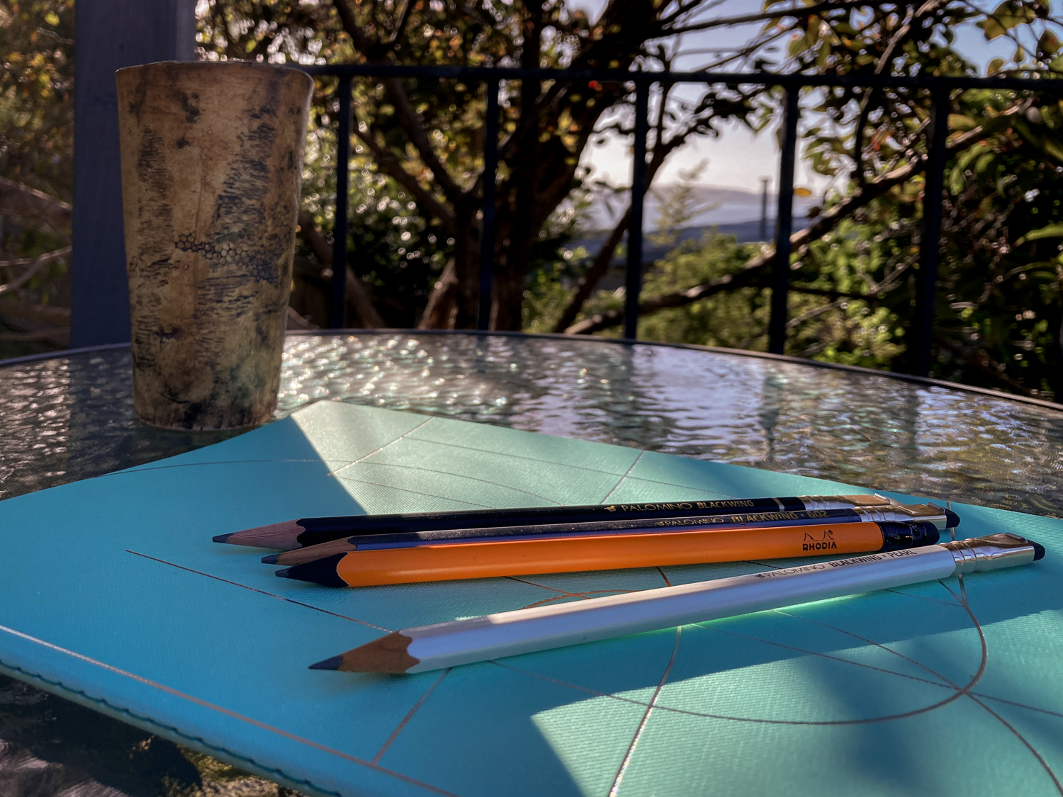 A green notebook on an outside table with four pencils on top of it. There is a coffee cup on the table