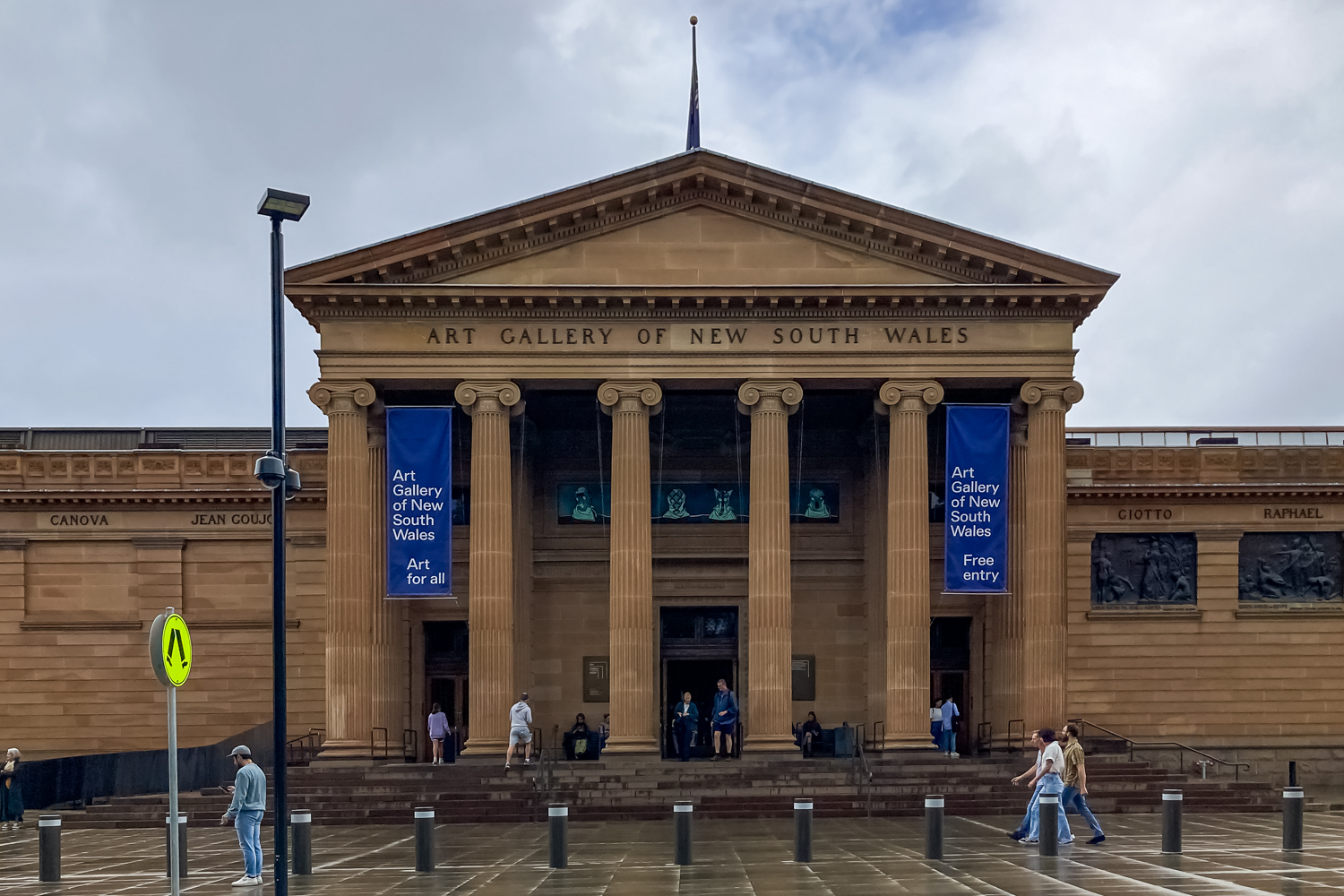 An old sandstone building with a triangle shaped roof and six columns at the entrance. The words Art Gallery of New South Wales on the front. Two blue signs either side of the entrance.