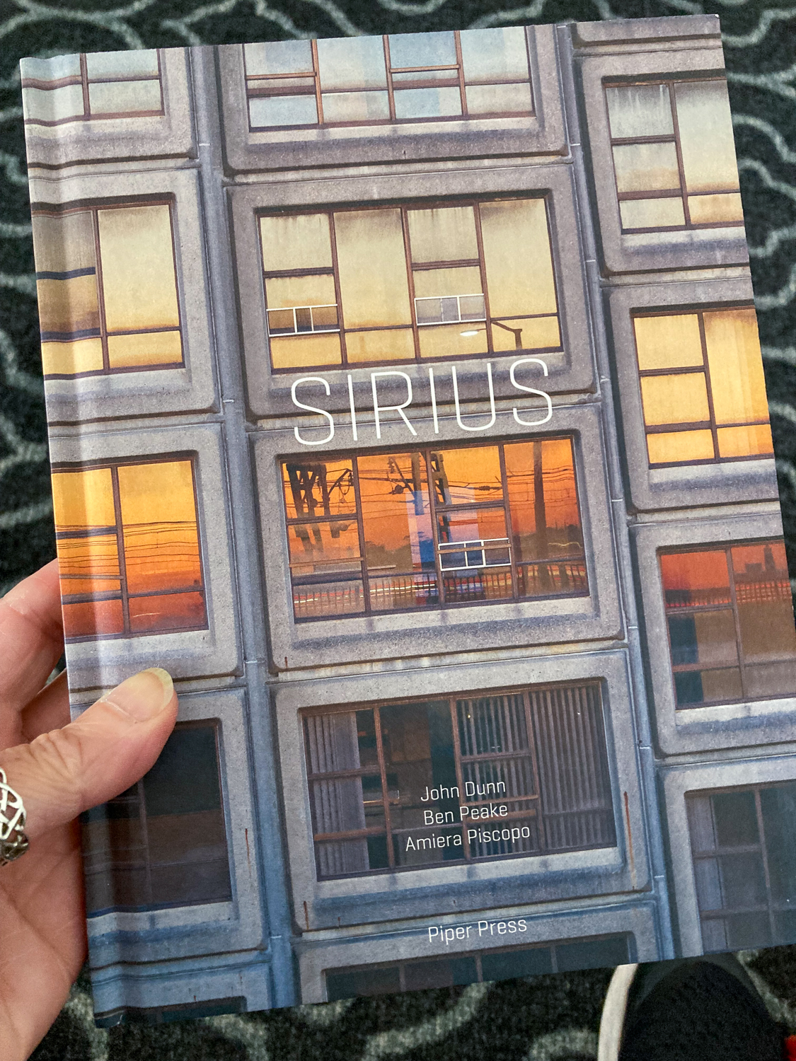 A hand holding a book with the title Sirius on the cover over a photo of a concrete building with orange, yellow and brown hues