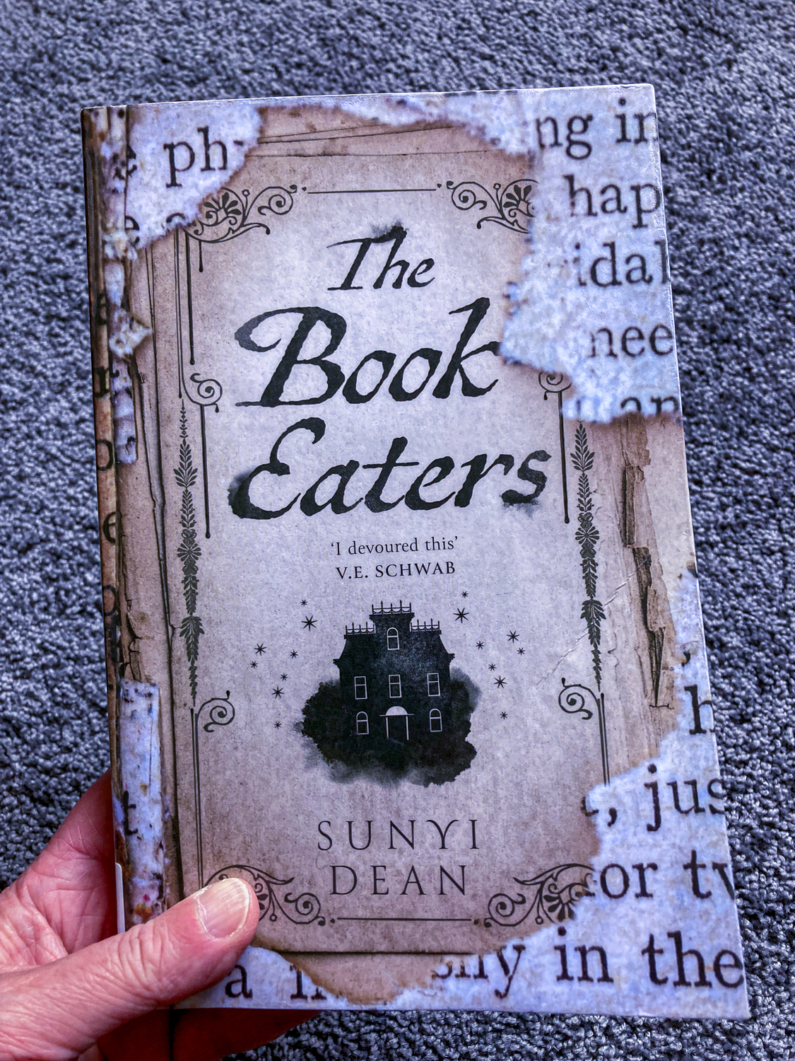 A hand holding a book, the cover featuring torn paper and the words The Book Eaters - Sunyi Dean