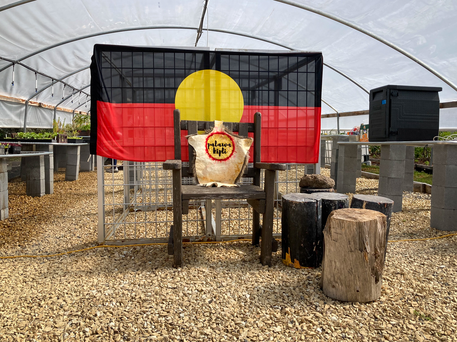 A woodenchair draped with an animal skin and a plaque with the words "palawa kipli" in front of an Aboriginal flag inside a poly tunnel