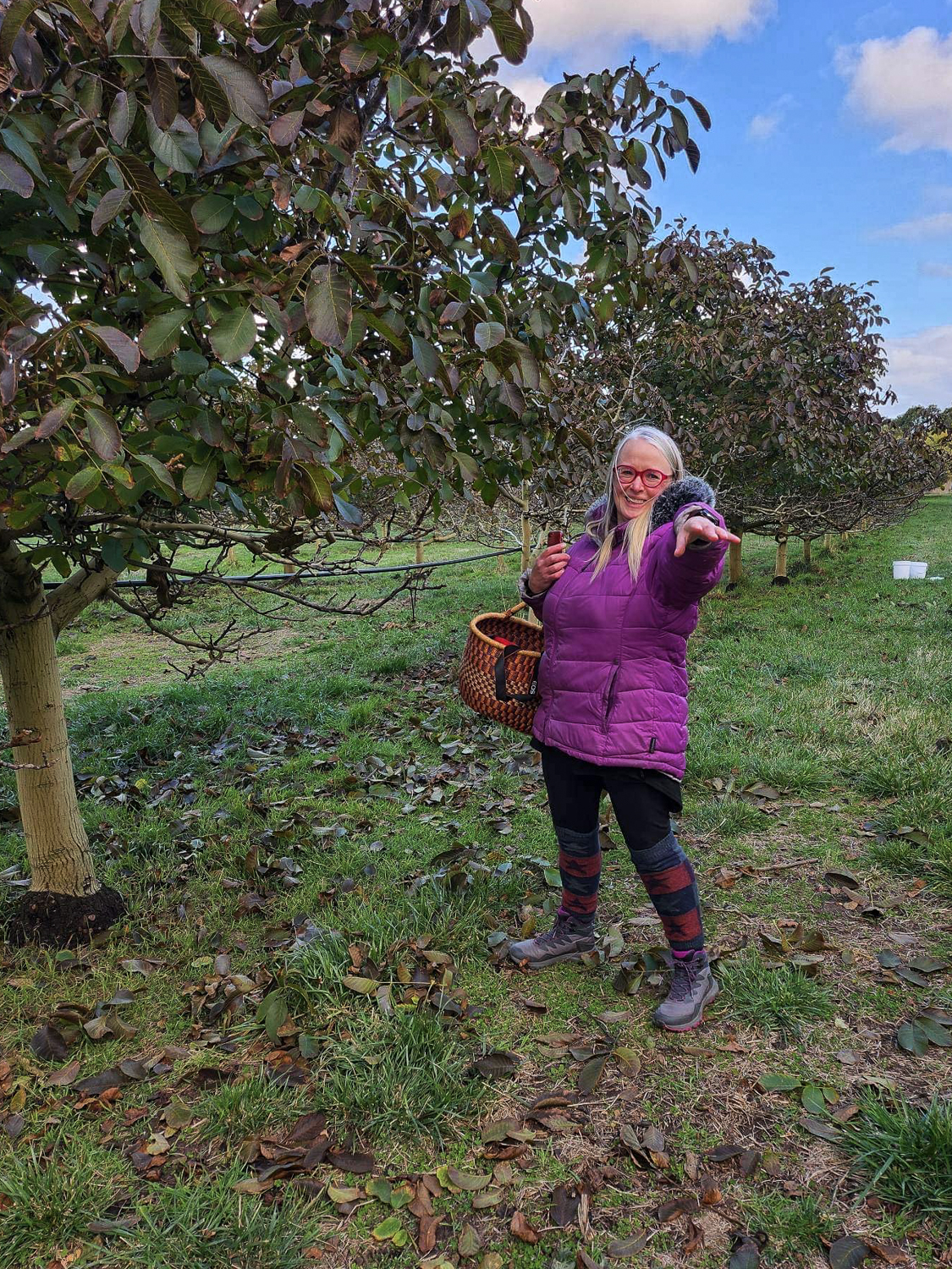 Barb is standing in the walnut orchard pointing at the camera. She is wearing a purple jacket and holding a large round basket
