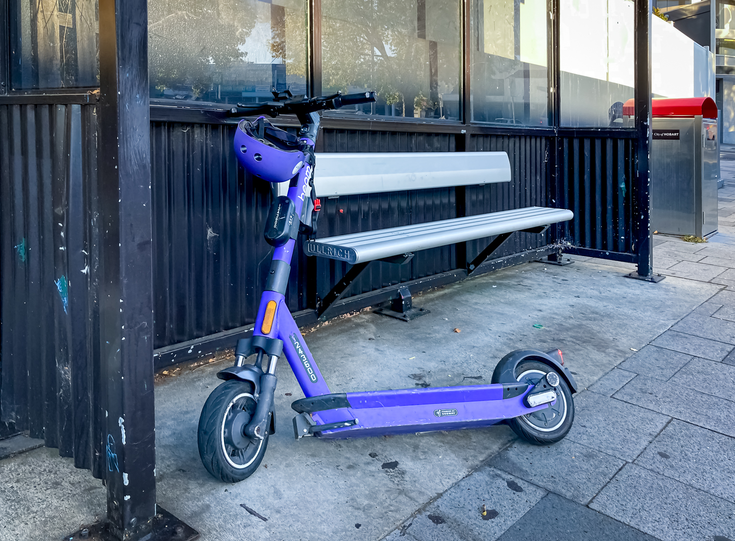 A purple e-scooter propped up against a seat under a bus shelter