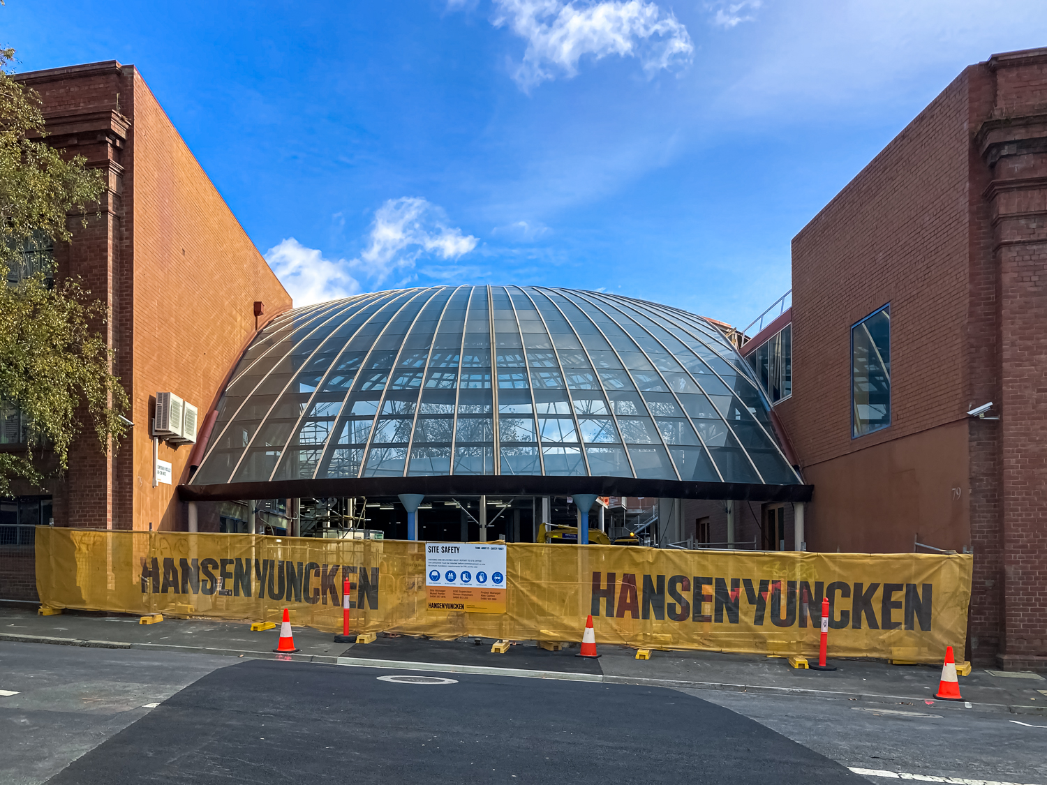 A dome shaped glass roof between two brick buildings, mostly shielded by a yellow fence with hoarding with the words Hansen Yuncken