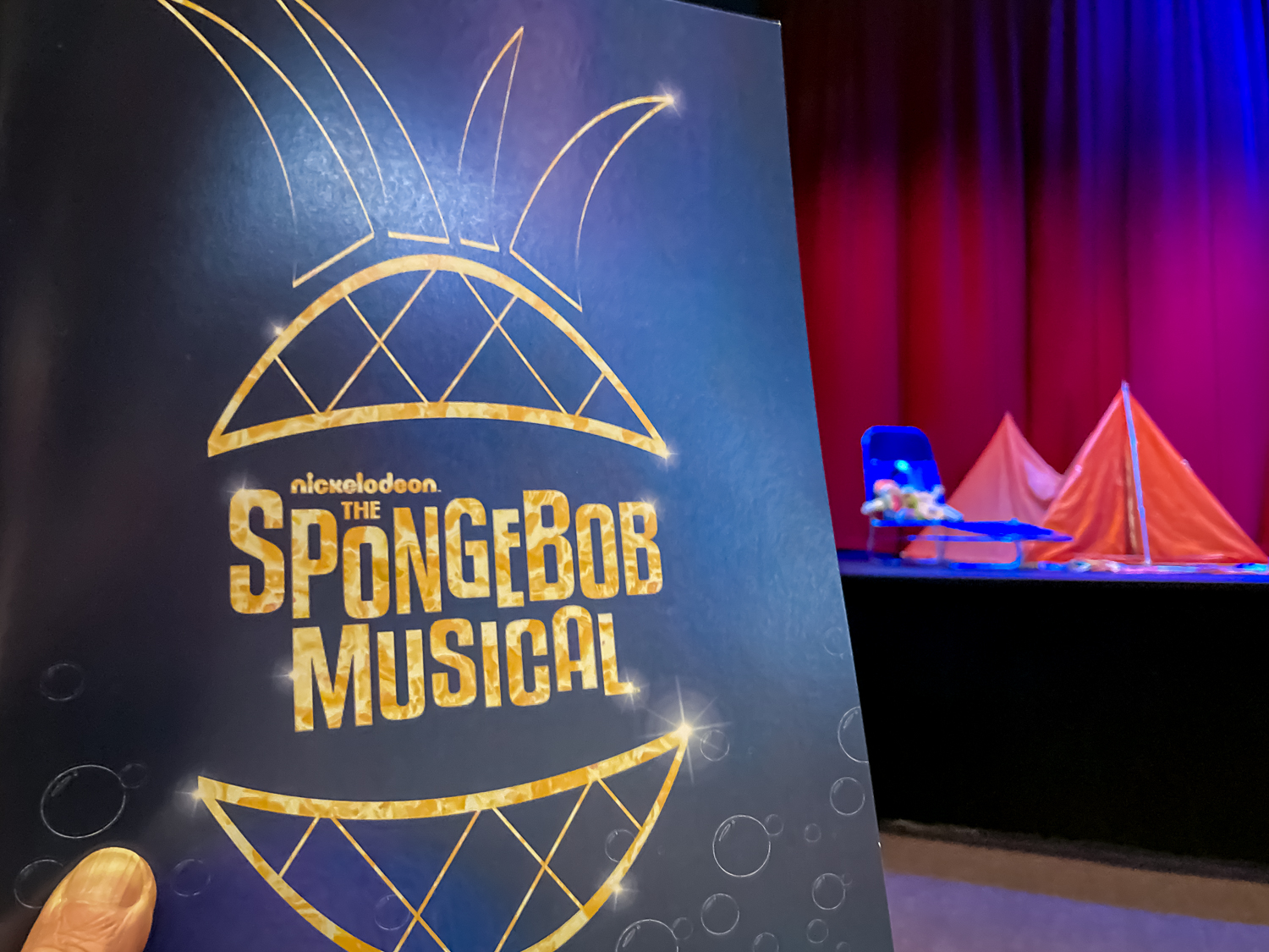 A blue page with the words SpongeBob Musical inside a pineapple, all in gold text, held up in front of a stage with red tents