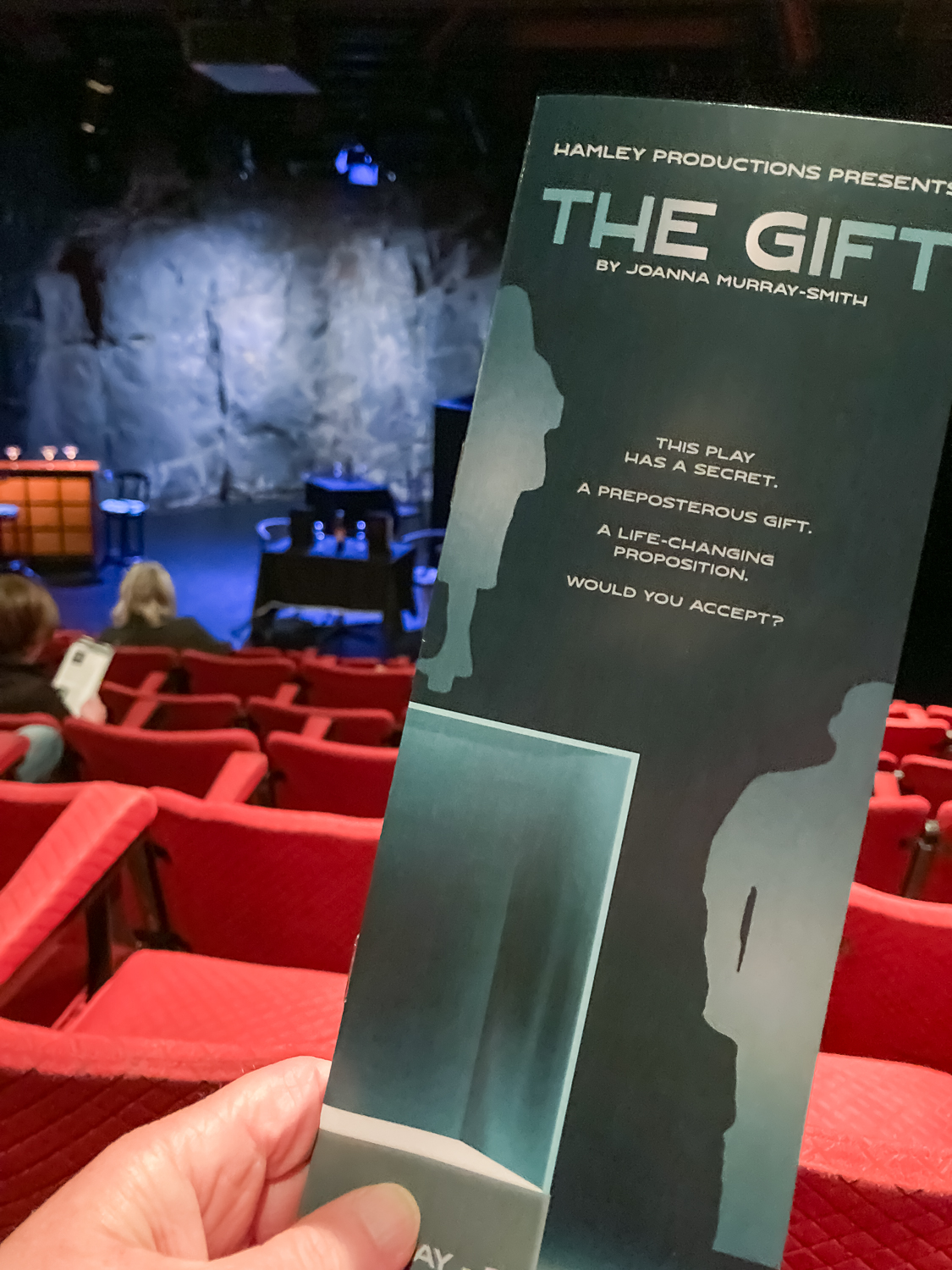 A long thin grey document with the words 'The Gift' held up in front of a theatre of red seats looking onto a stage