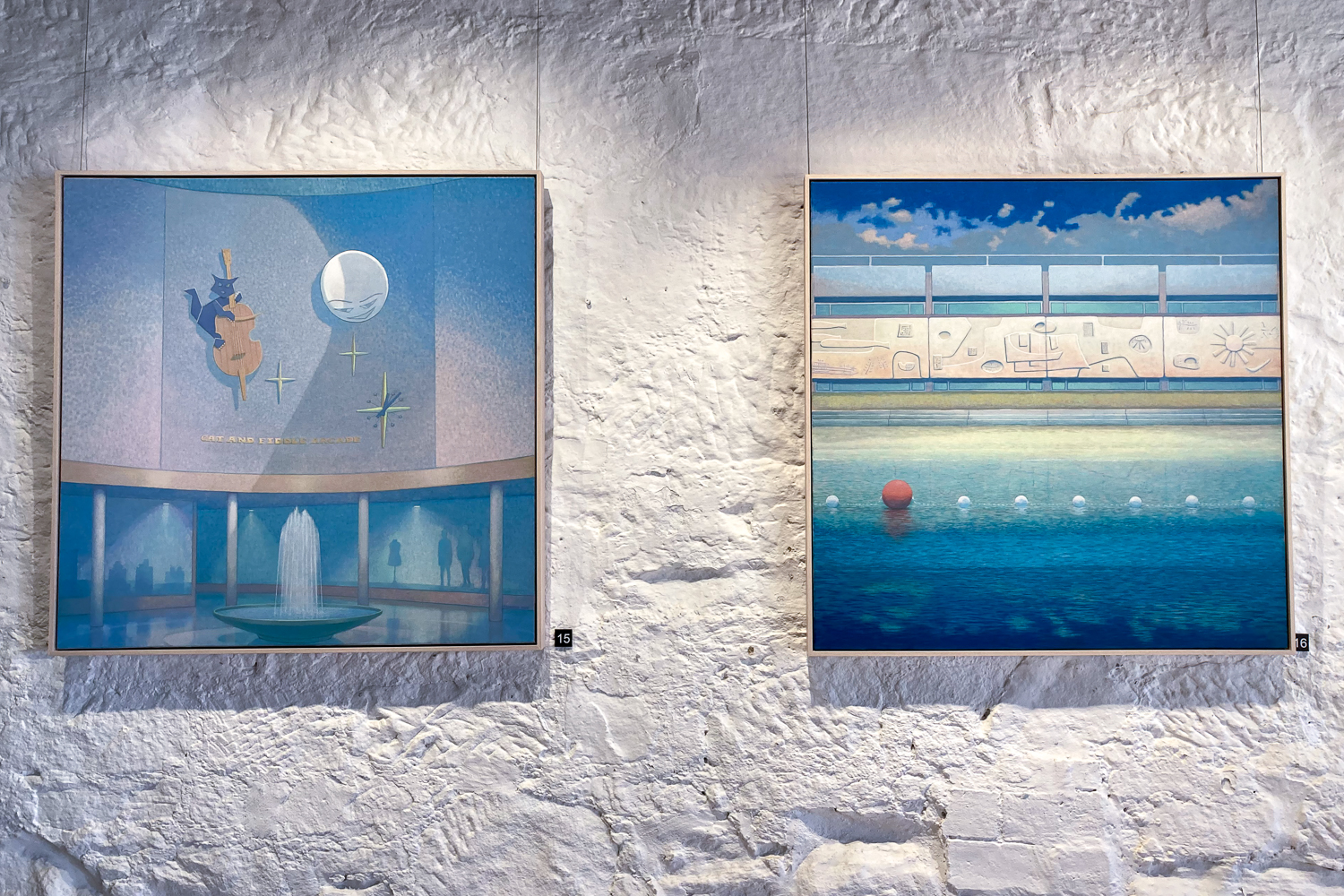 Two paintings on a white textured gallery wall. One is of a moon and a cat with a fiddle, the other a sculptured bathing pavilion on the beachfront
