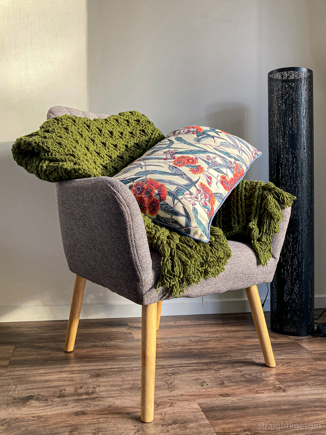 A grey chair with a green throw rug and a long cushion with a Tasmanian native plant design