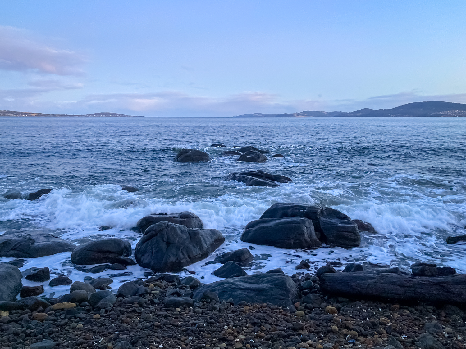 Water breaking over small rocks on a rocky shoreline, with low clouds over the horizon