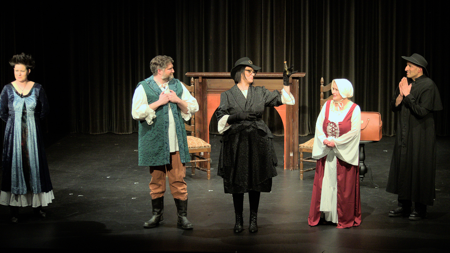 Five actors dressed in medieval costumes on a theatre stage. The character in the middle, dressed in black, is holding something up for the others to look at