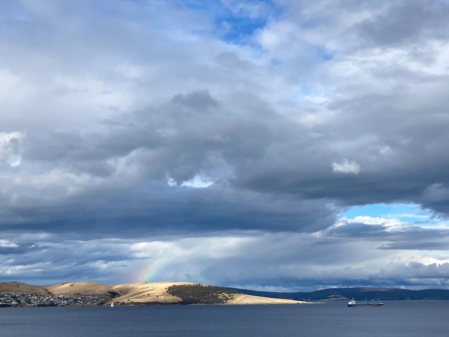 A small land mass across the river with a partial rainbow and heavy cloud cover