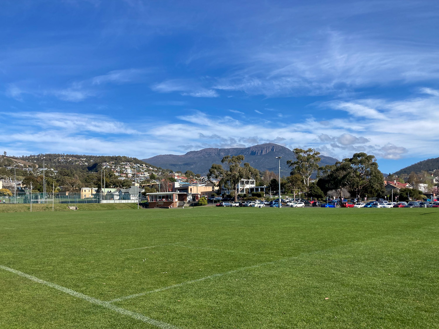 A sports oval with some university buildings in the background, and further back, kunanyi/Mt Wellington. The sky is blue with some fluffy clouds