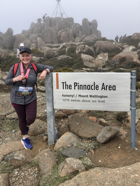 Barb is standing in front of some rocks leaning on a sign that says "The Pinnacle Area". She is wearing sports clothes, a cap and has a medal around her neck. She is smiling.