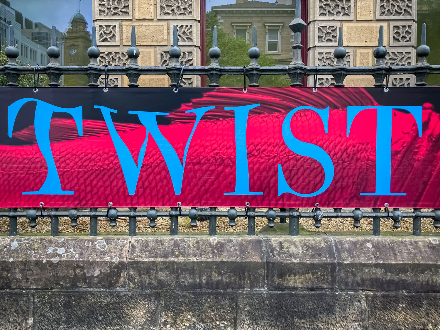The word "TWIST" in blue text on a pink background of a sign hanging on a wrought iron fence outside a sandstone building