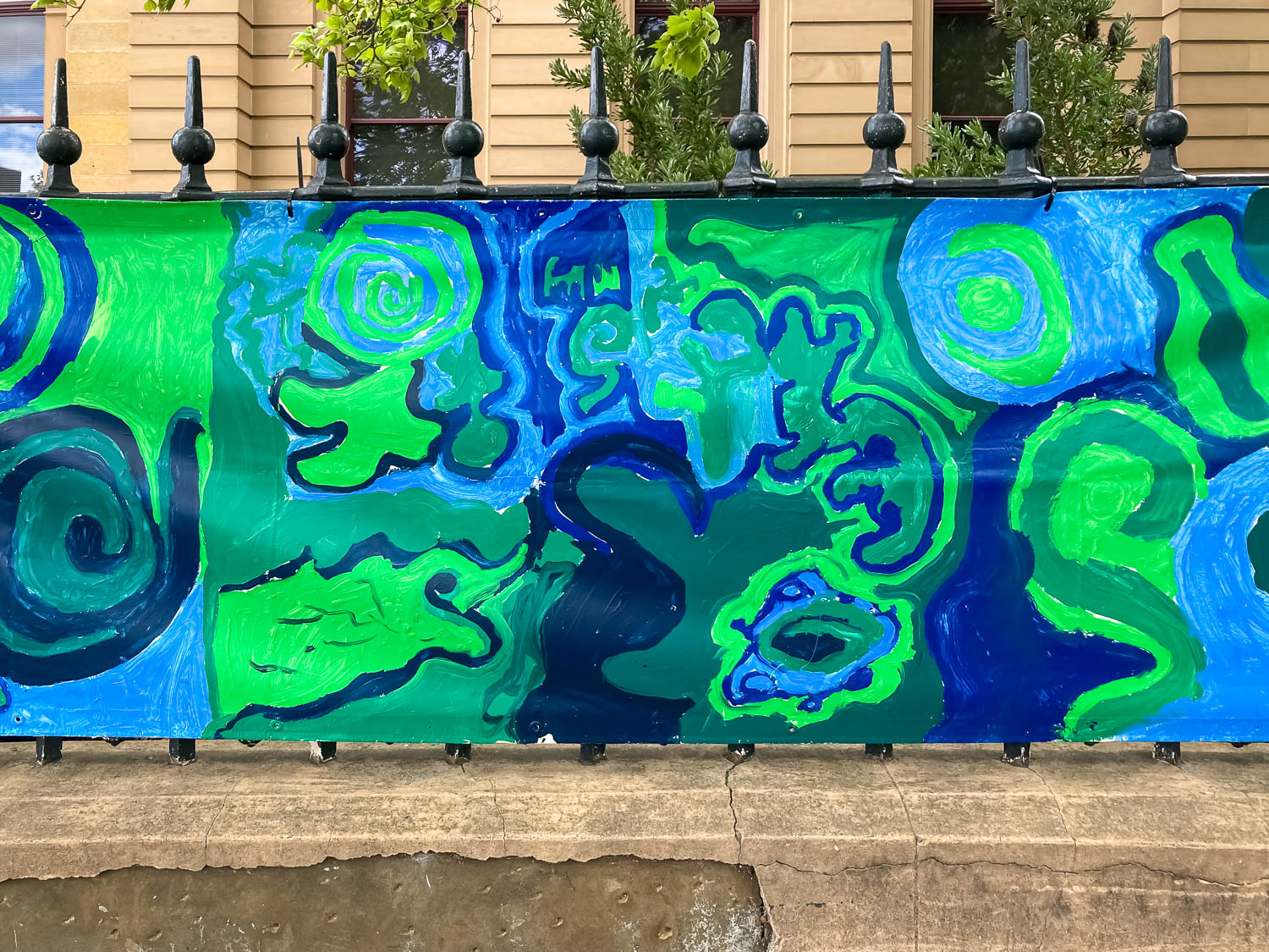 A green and blue swirling artwork on a banner on a wrought iron fence outside a sandstone building