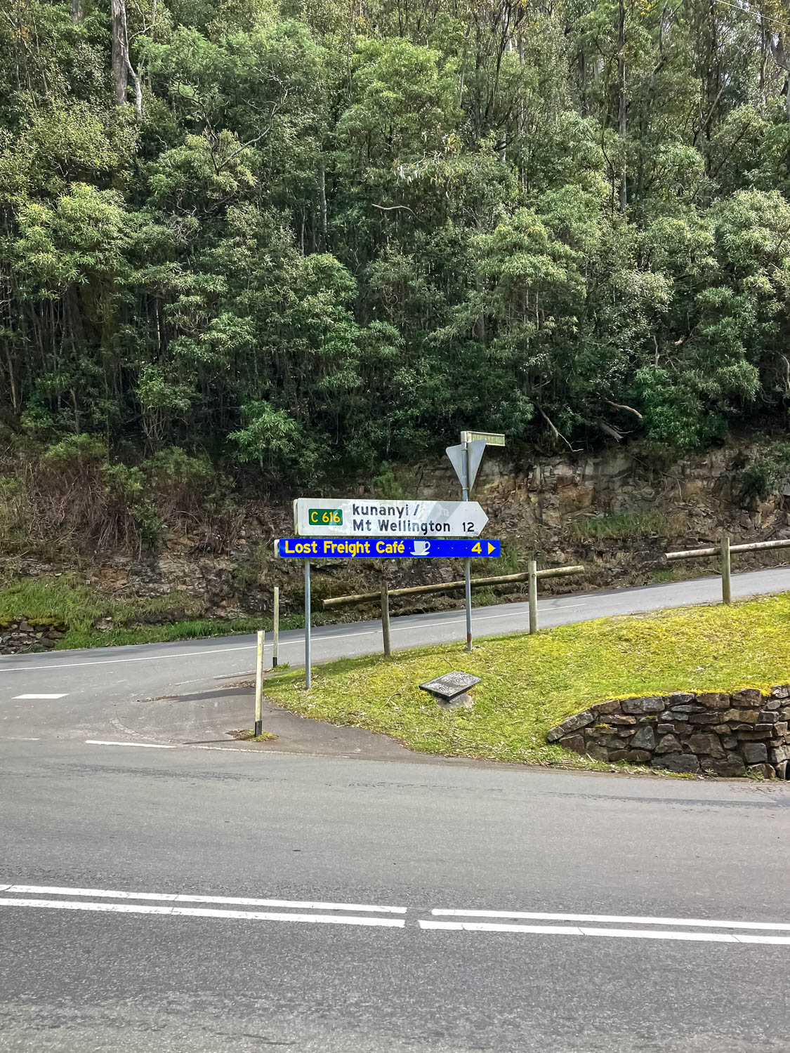 A junction of two roads, one going back to the right with a sign pointing to kunanyi/Mt Wellington 12 km. There are trees in the background