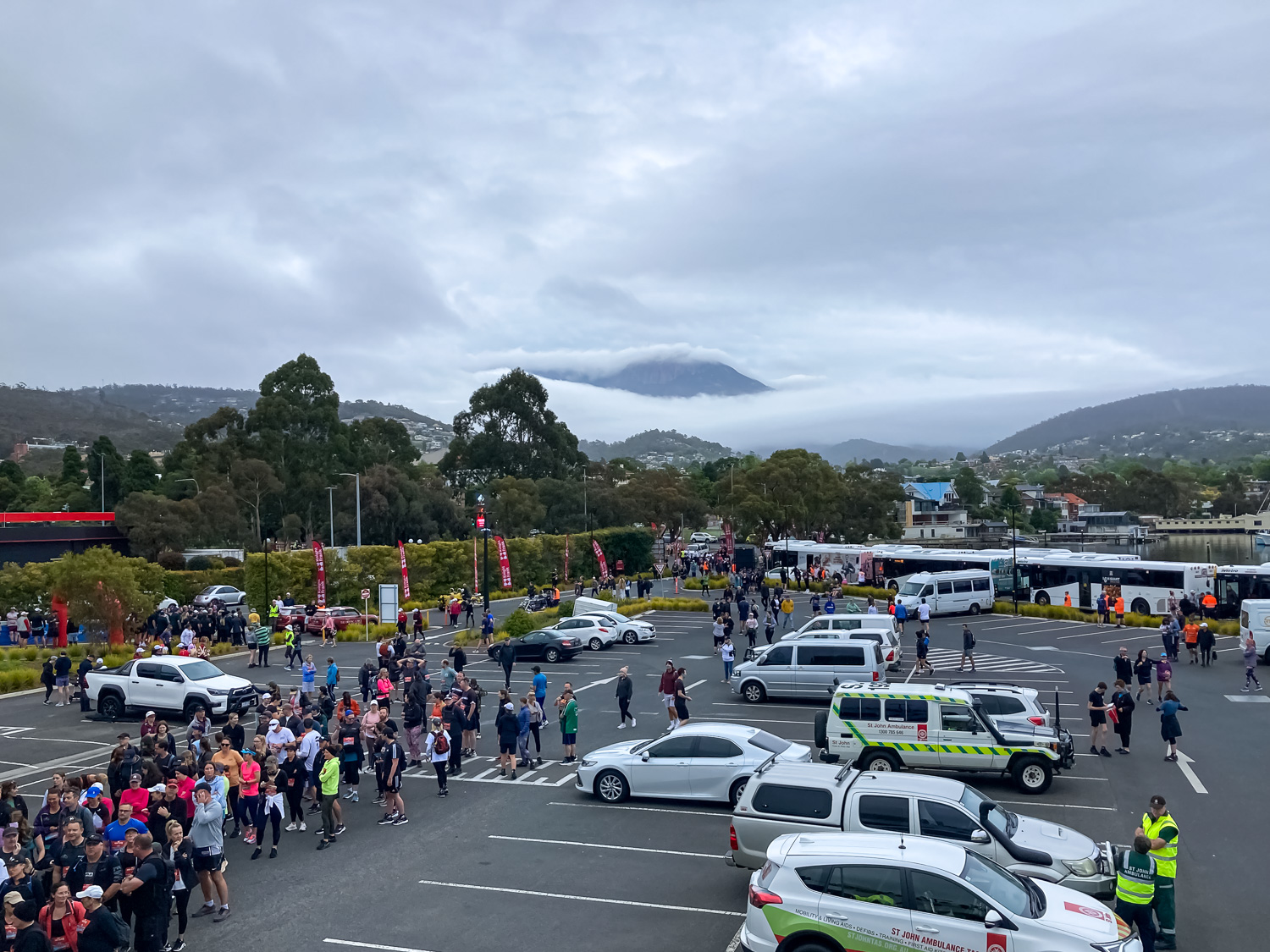 A car park filled with people, emergency vehicles and buses. In the background is a cloud covered mountain