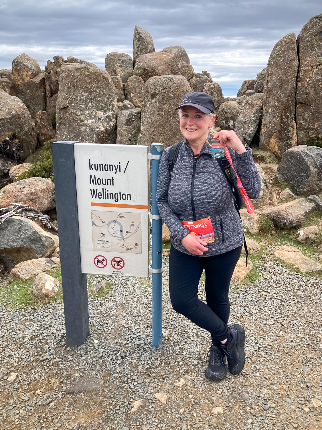 A woman wearing a black cap, a grey jacket and black pants standing in front of rocks and a sign that reads kunanyi/Mount Wellington