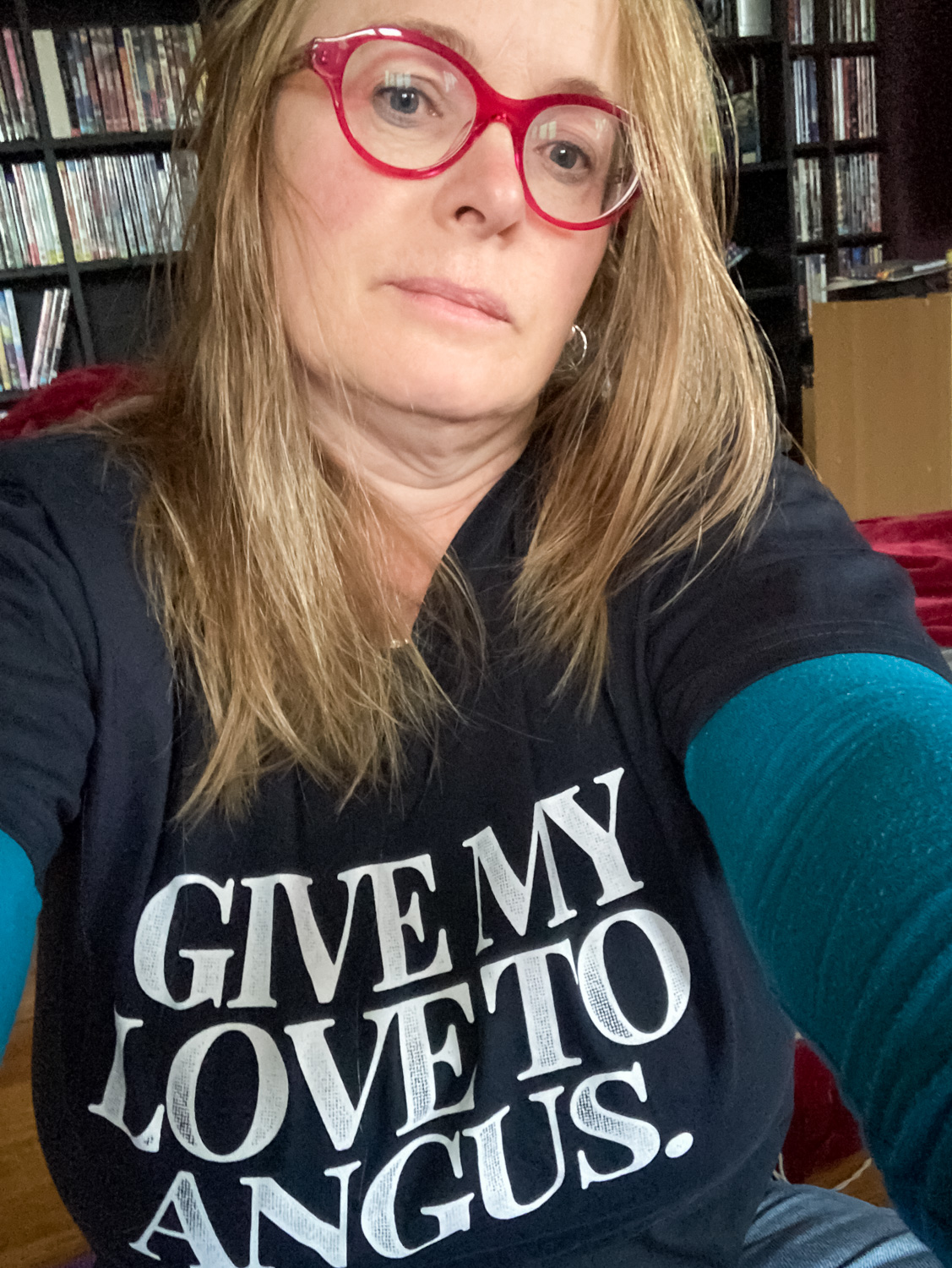 Barb is wearing a dark blue t-shirt with the words "GIVE MY LOVE TO ANGUS" o the front. She has long blonde hair and pink glasses