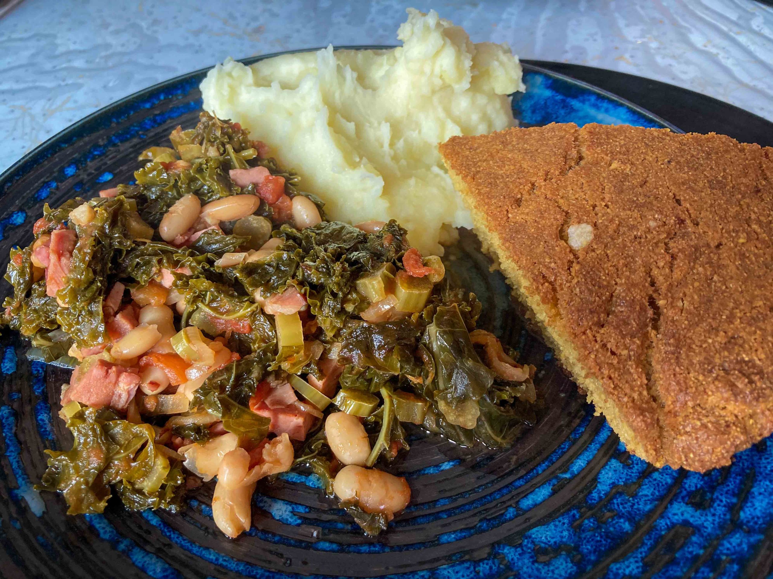 A blue plate with a serving of cooked kale, white beans and ham served with potato mash and cornbread