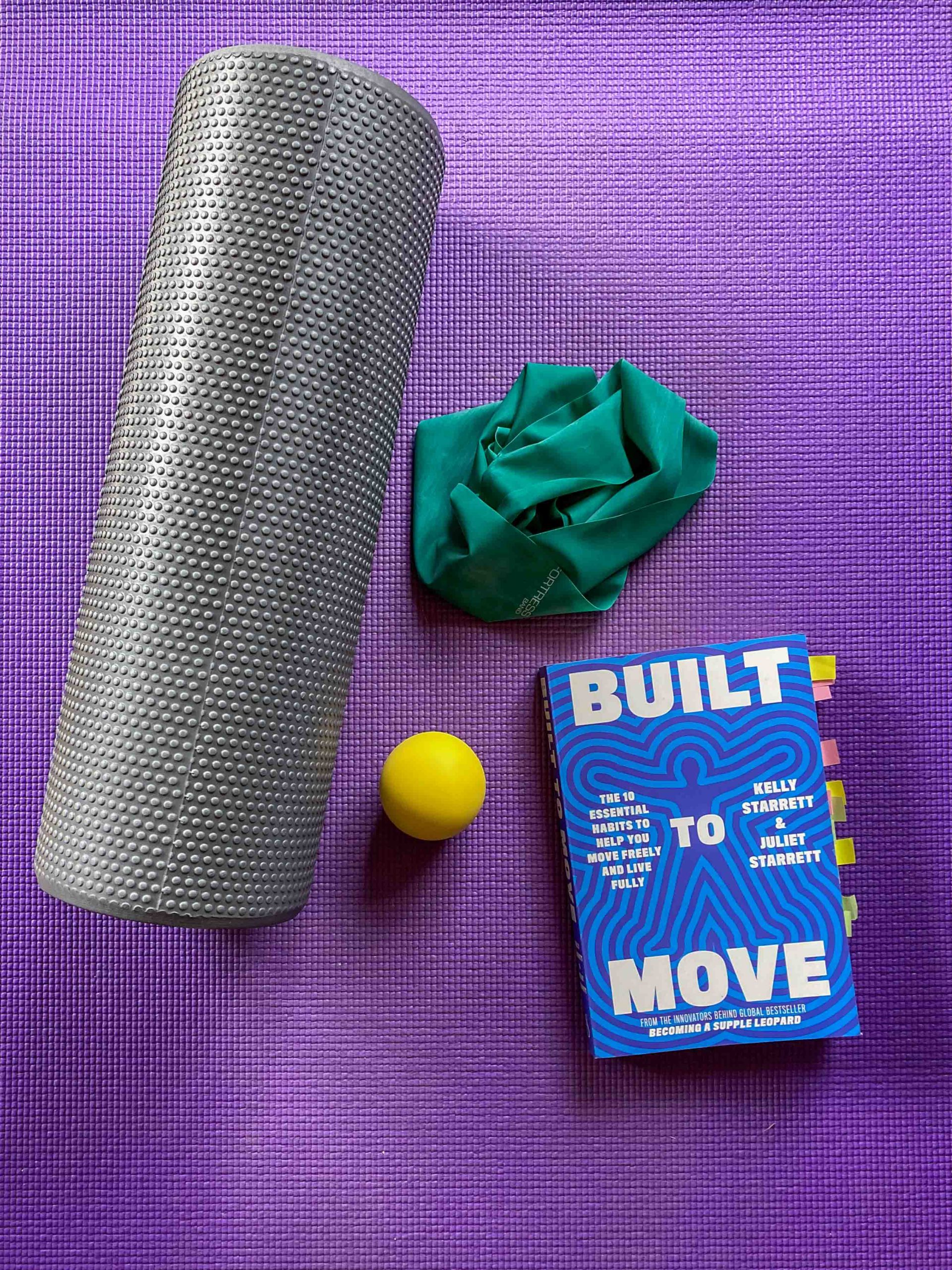 A purple yoga mat with a grey foam roller, a green stretchy band, a small yellow ball and a copy of the book Built to Move