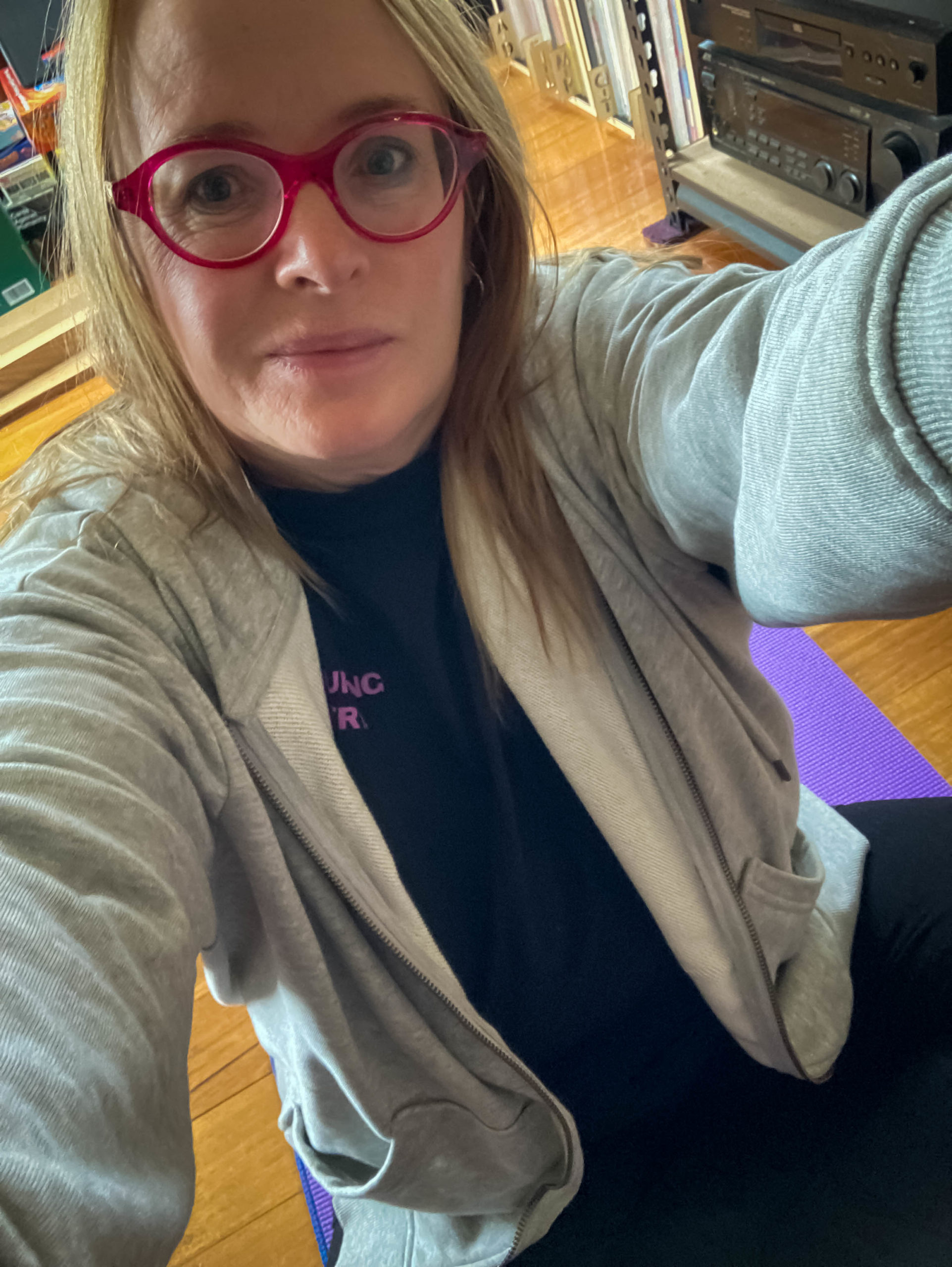 Barb is wearing a grey hoody and pink glasses. She is attempting to stand up from a seated position on the floor without using her hands