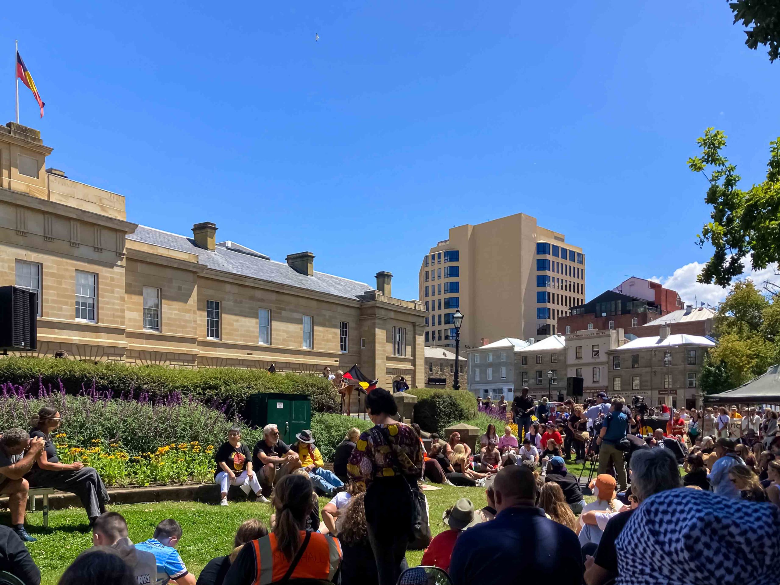 A gathering of people on the lawns outside a large sandstone building. The Aboriginal flag is flying