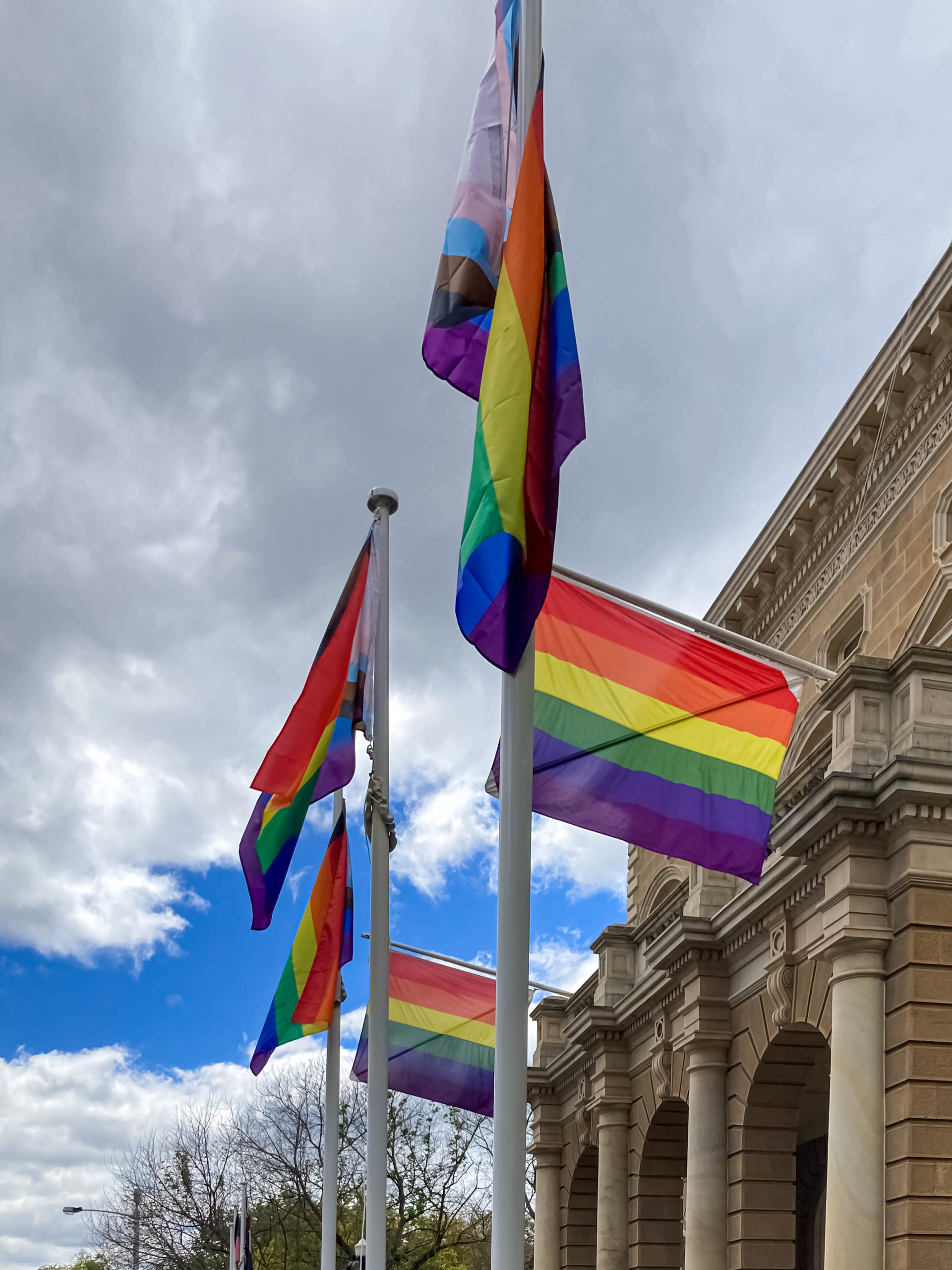 Five rainbow flags representing the LGBTIQA+ community flying outside a large sandstone building