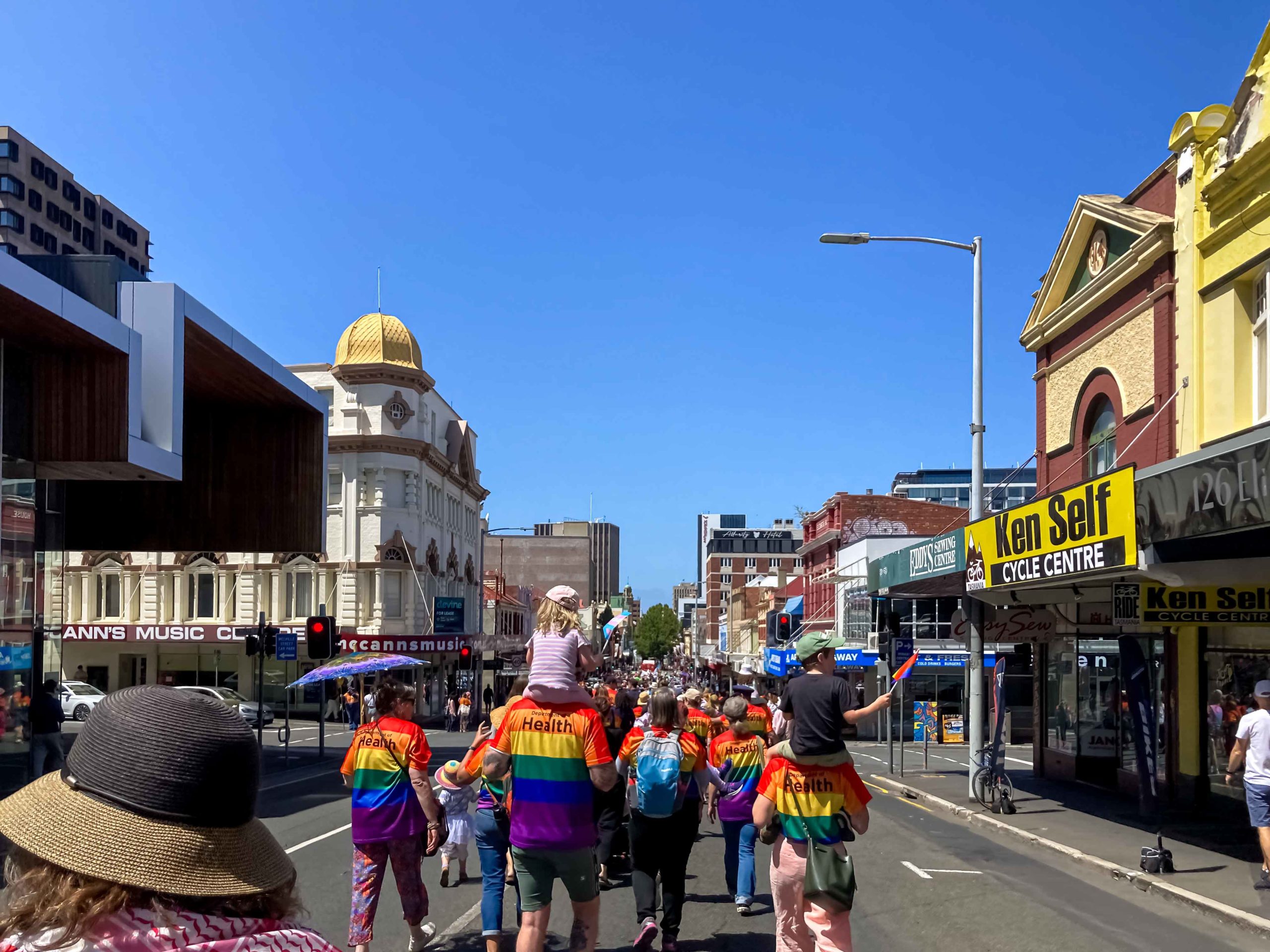A back view of people wearing rainbow shirts walking through the city streets as part of a large parade