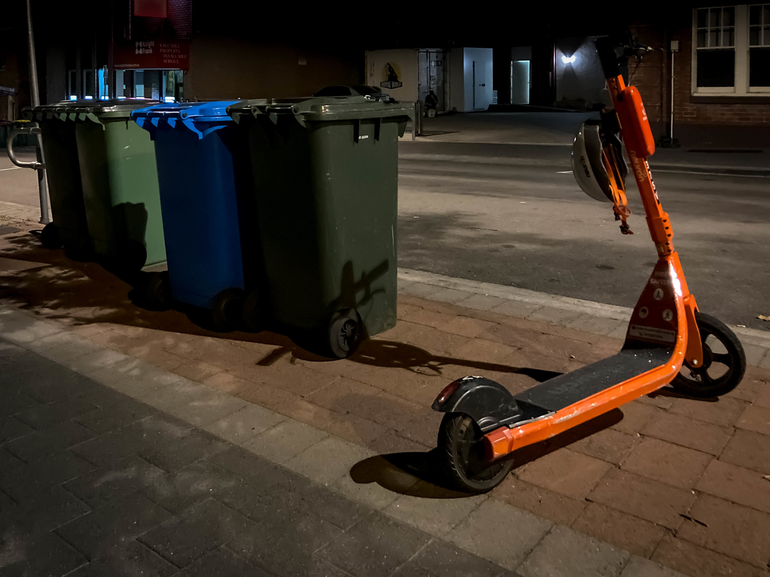 An orange e-scooter parked next to a row of bins in a dark street