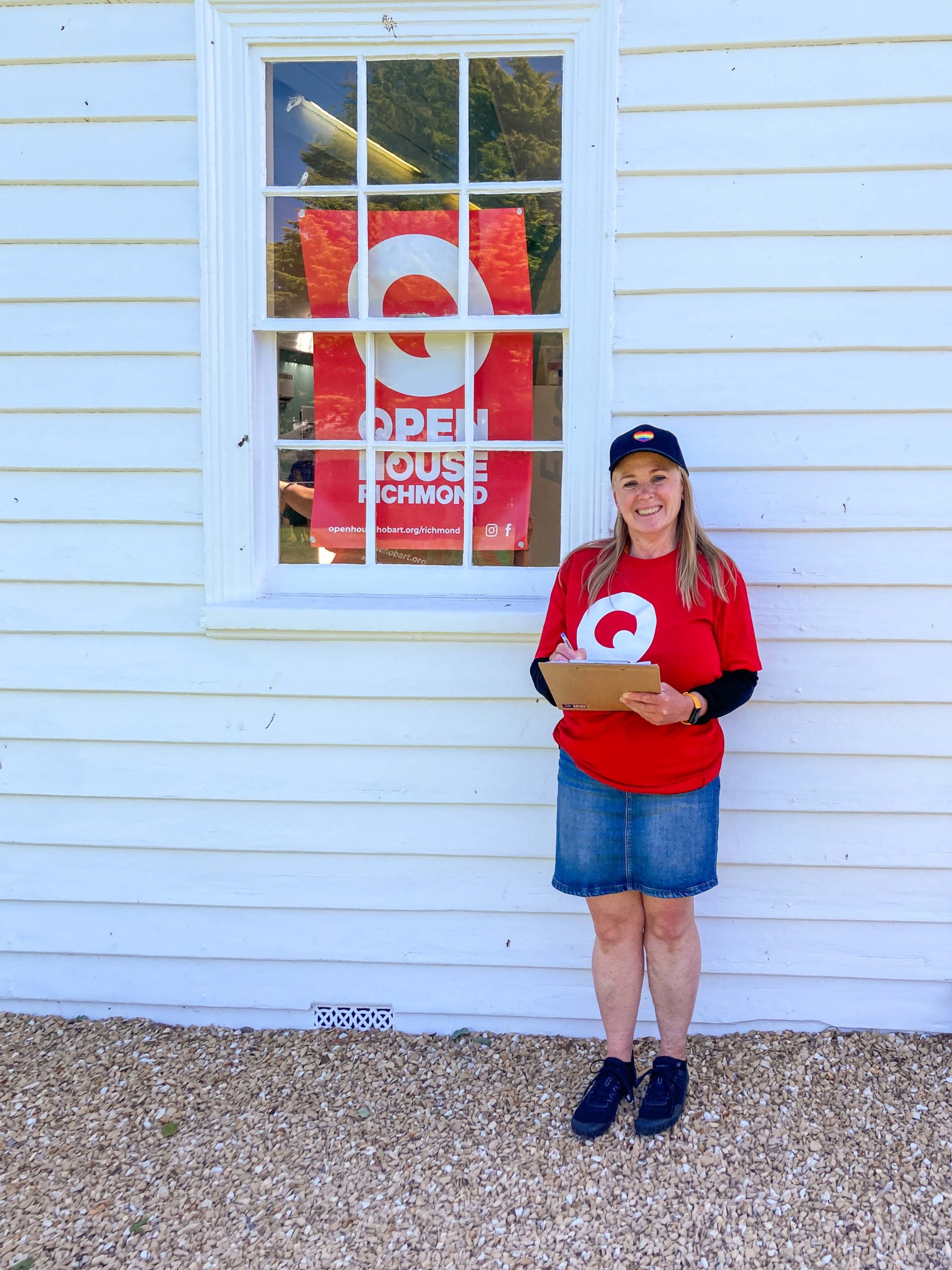 Barb is standing outside a white wooden building with a red poster bearing the words 'Open House Richmond'. Barb is wearing a red t-shirt, a blue skirt, a black cap and she is holding a clipboard