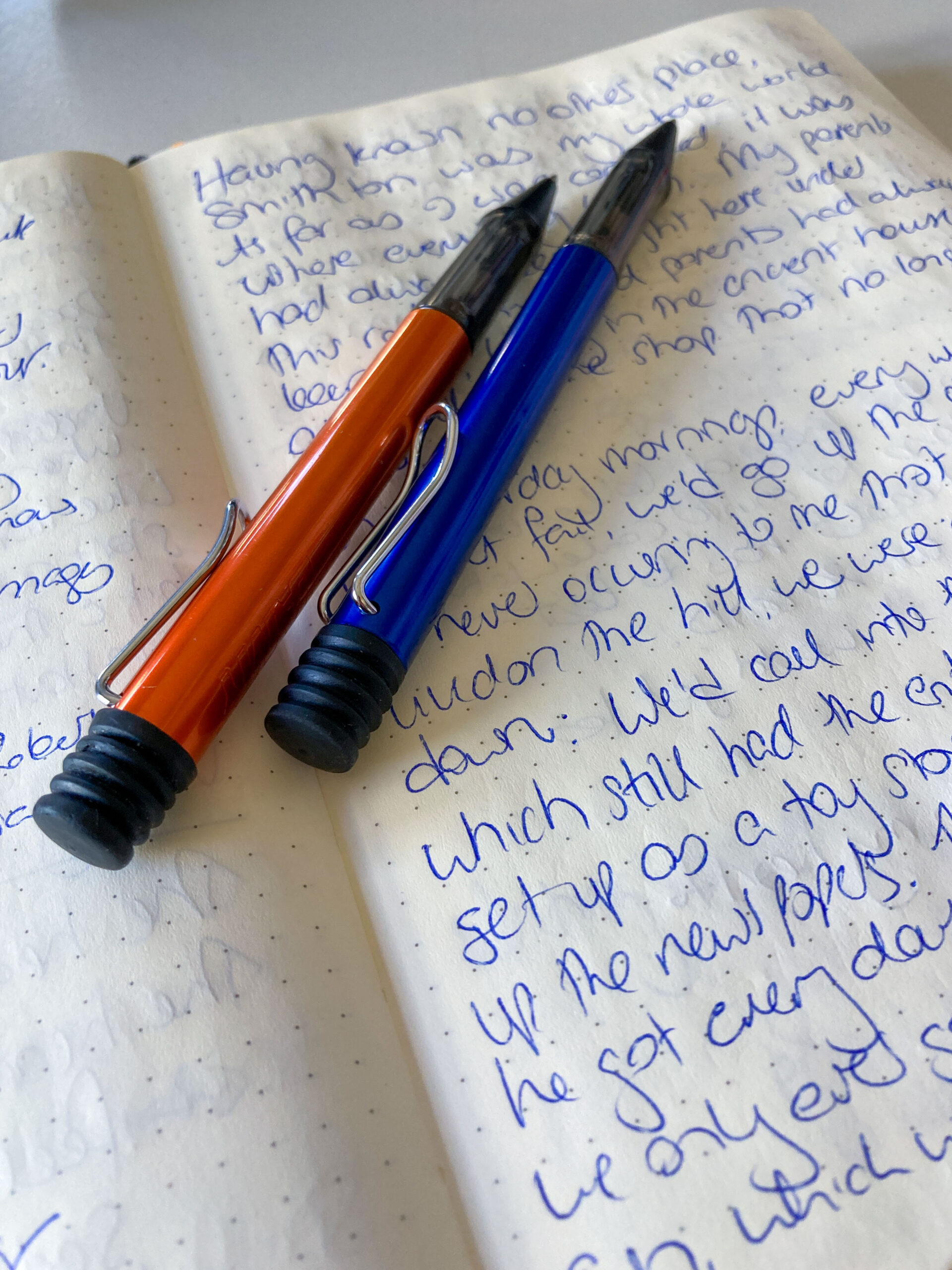 A page of handwriting in blue ink with orange and blue pens resting on the paper