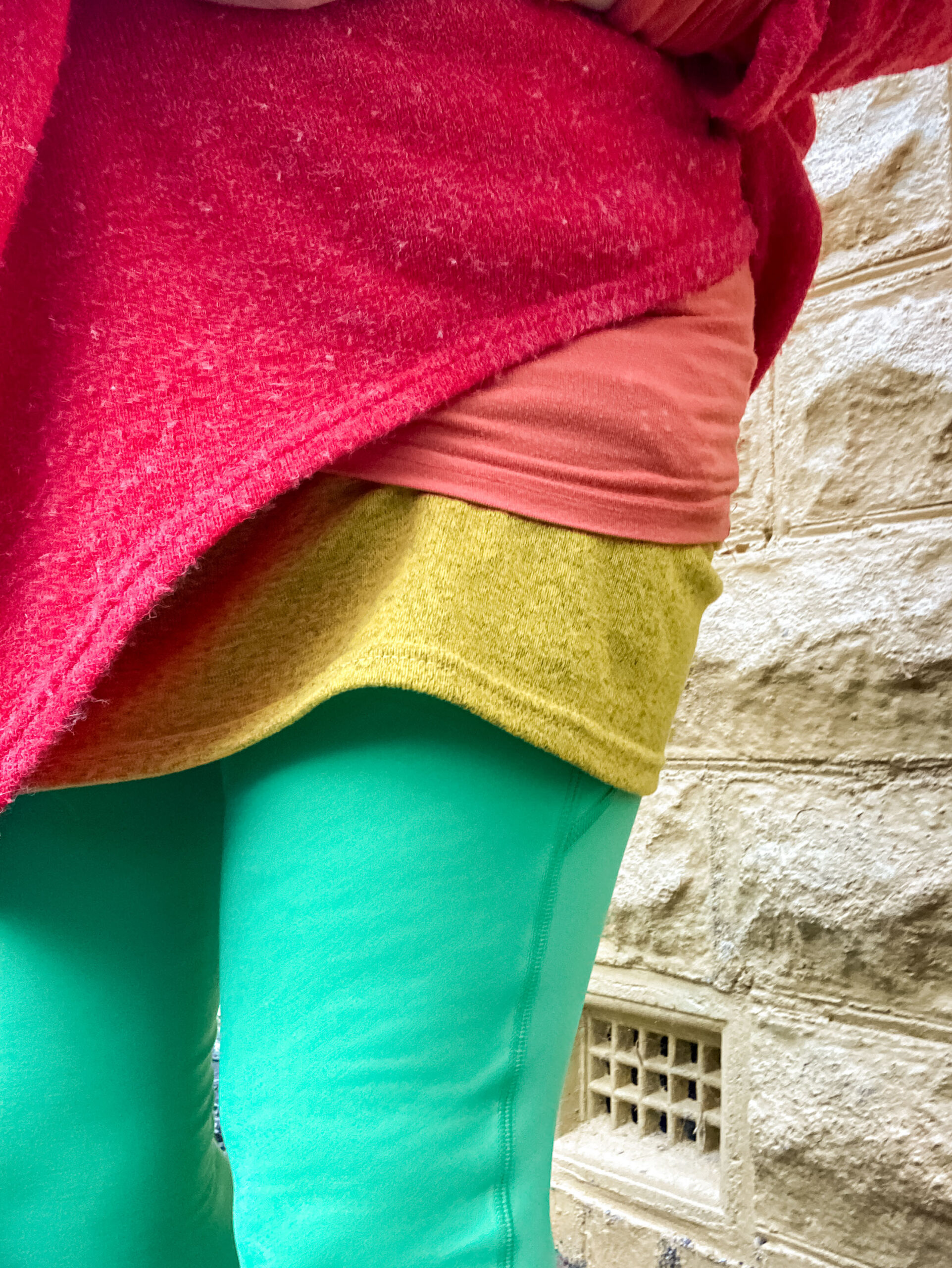 A close up of a person wearing green leggings, yellow skirt, orange top and a red jumper. The image doesn't show their face, just their upper leg and abdomen