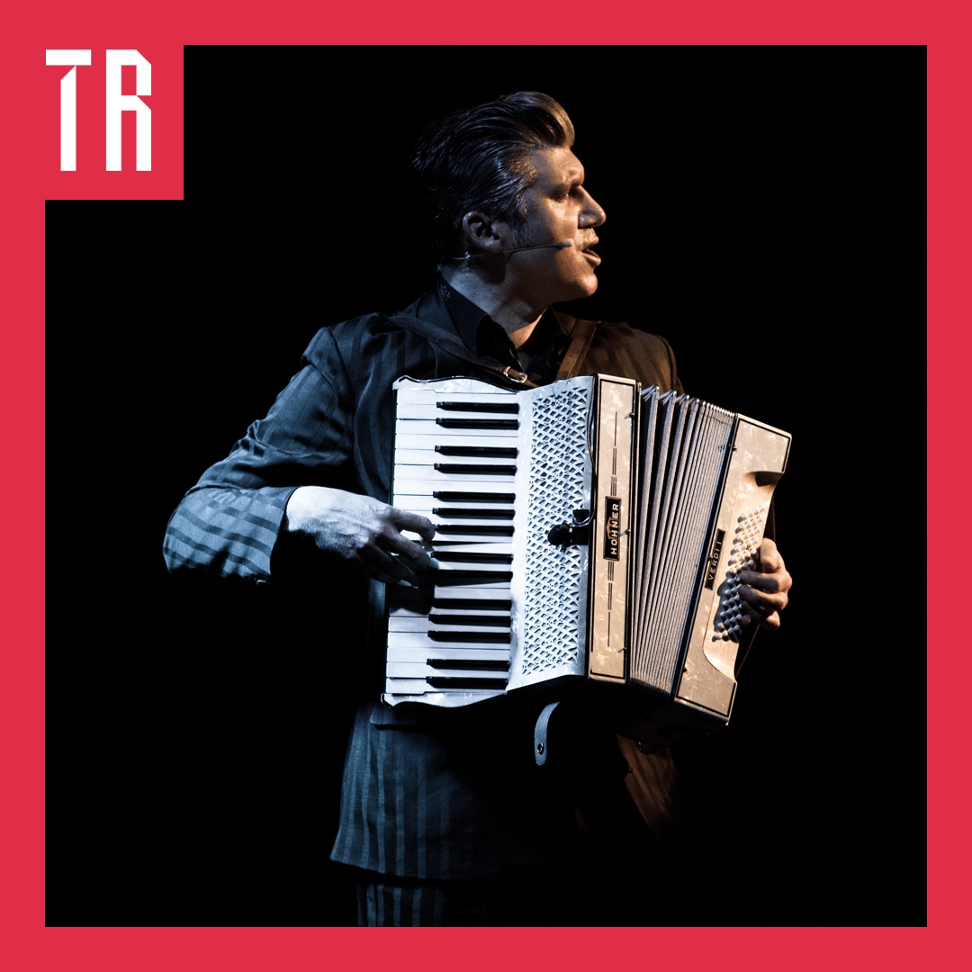 A man faces the right of the photo. He is dressed in a suit playing an accordion. The image has a red border and the letters TR in the top left hand corner