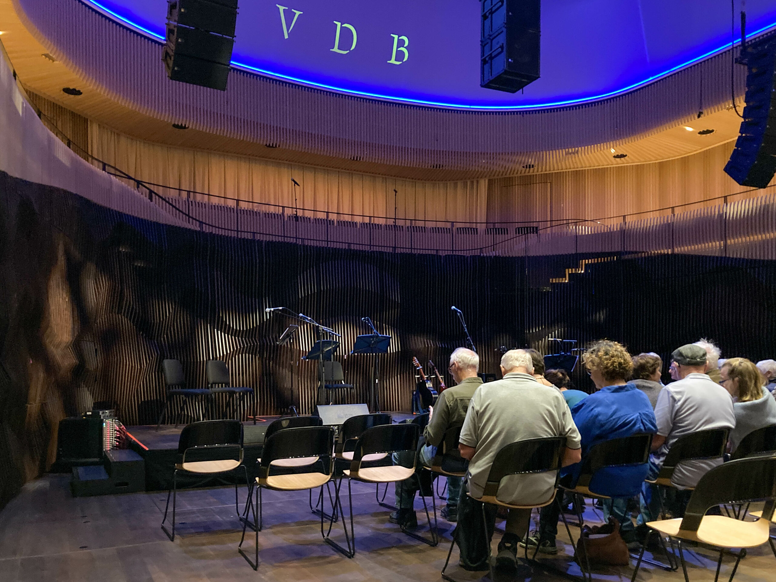 People sitting on chairs facing a stage set up for six musicians. The letters VBD appear in purple lighting above the stage