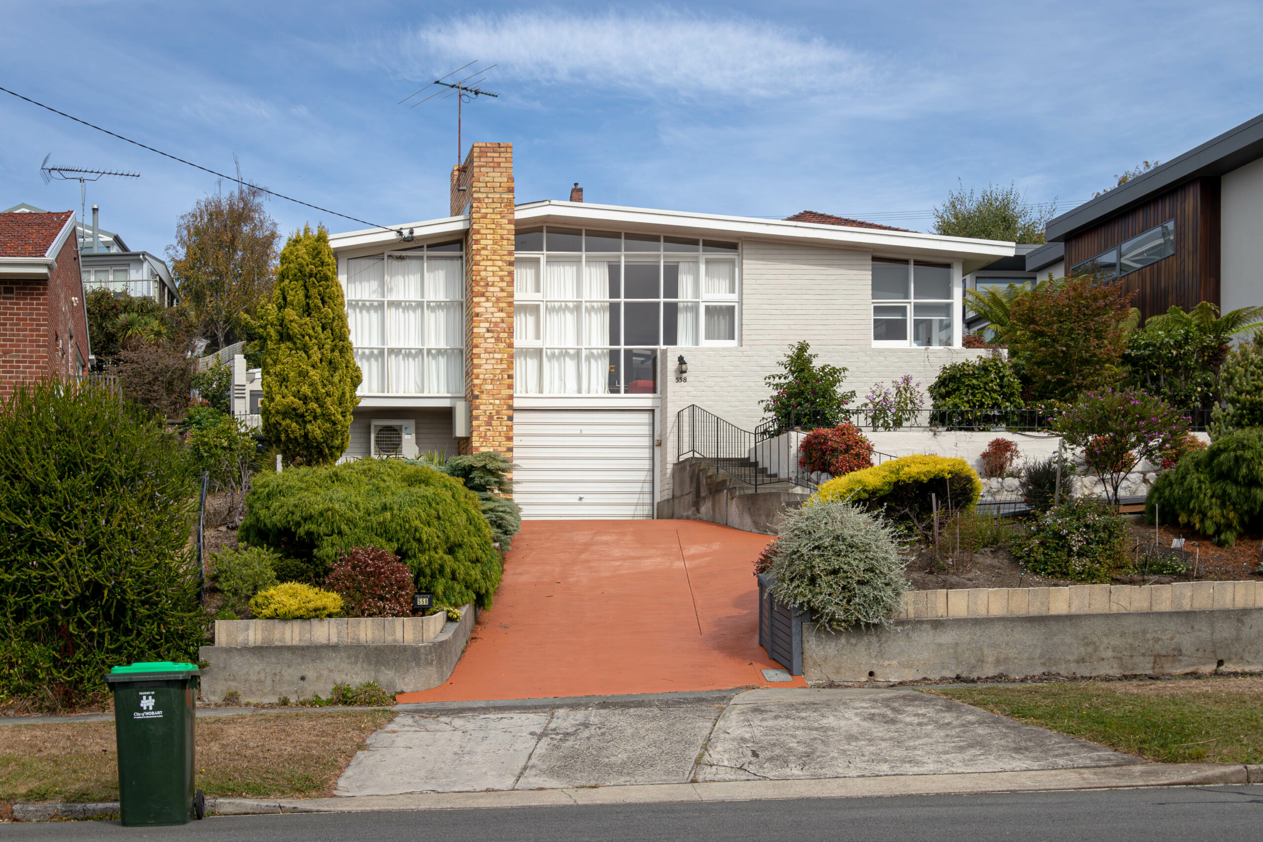 A mid-20th century brink house painted white, with a grid of small square windows on the front wall and an orange brick chimney. There is an orange driveway leading to a garage door