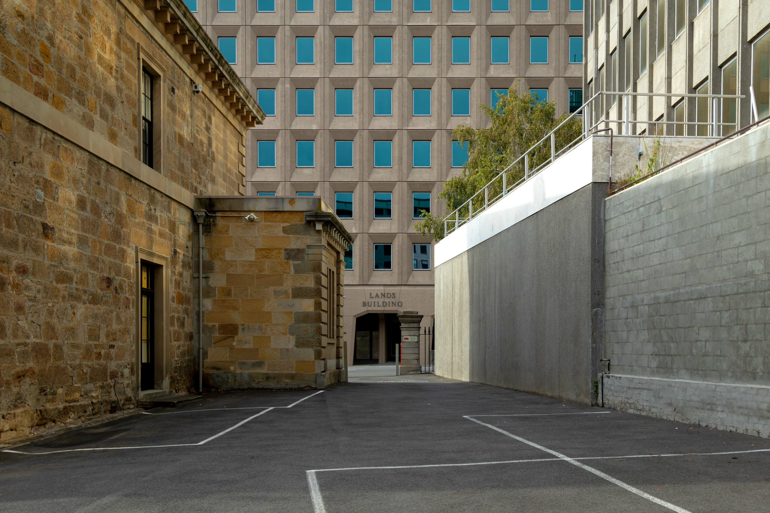 a large concrete building with rows of identical windows, seen in between an old sandstone building and a concrete wall