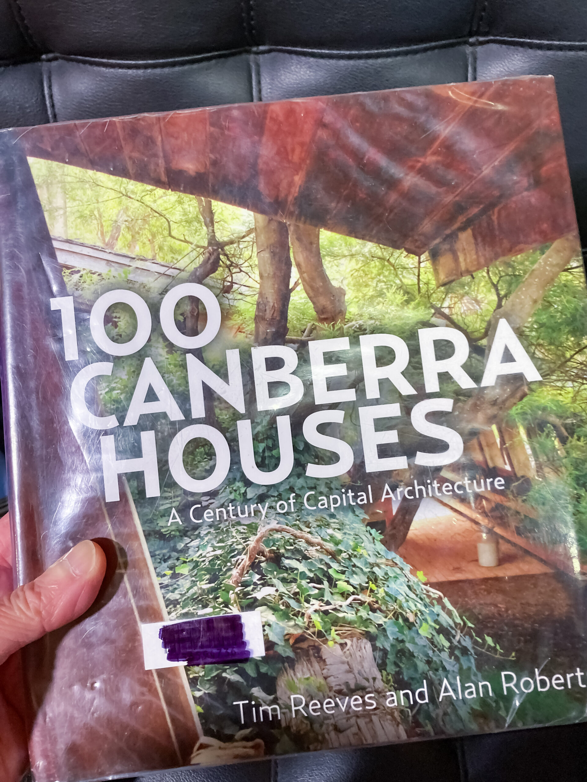 The cover of a book titled 100 Canberra Houses which has a picture of a timber structure surrounded by green plants