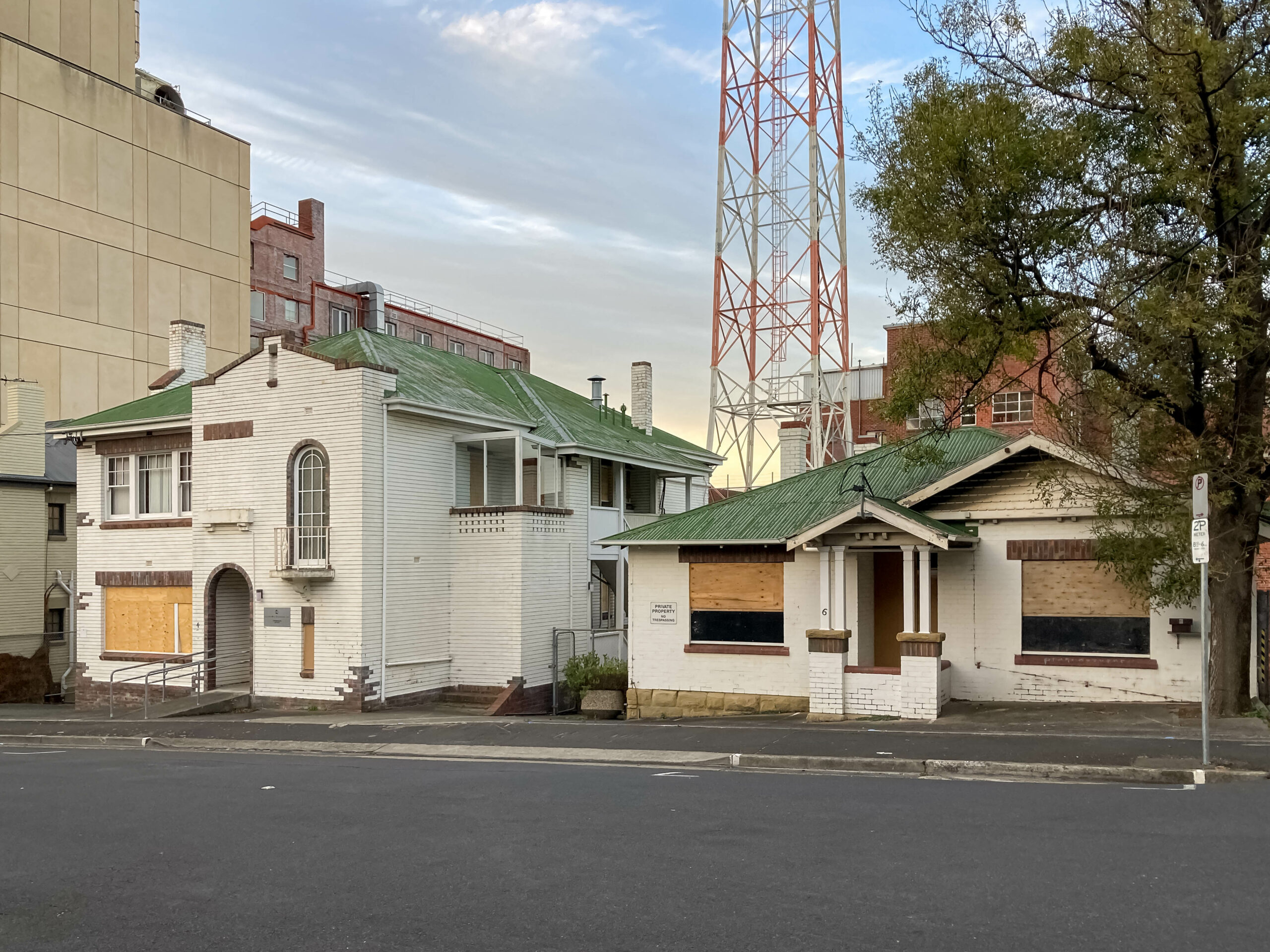 Two mid-20th century white houses with boarded up windows. They both have battered green roofs. The one on the left is weatherboard and is two storeys. The one on the right is single-storey brick.