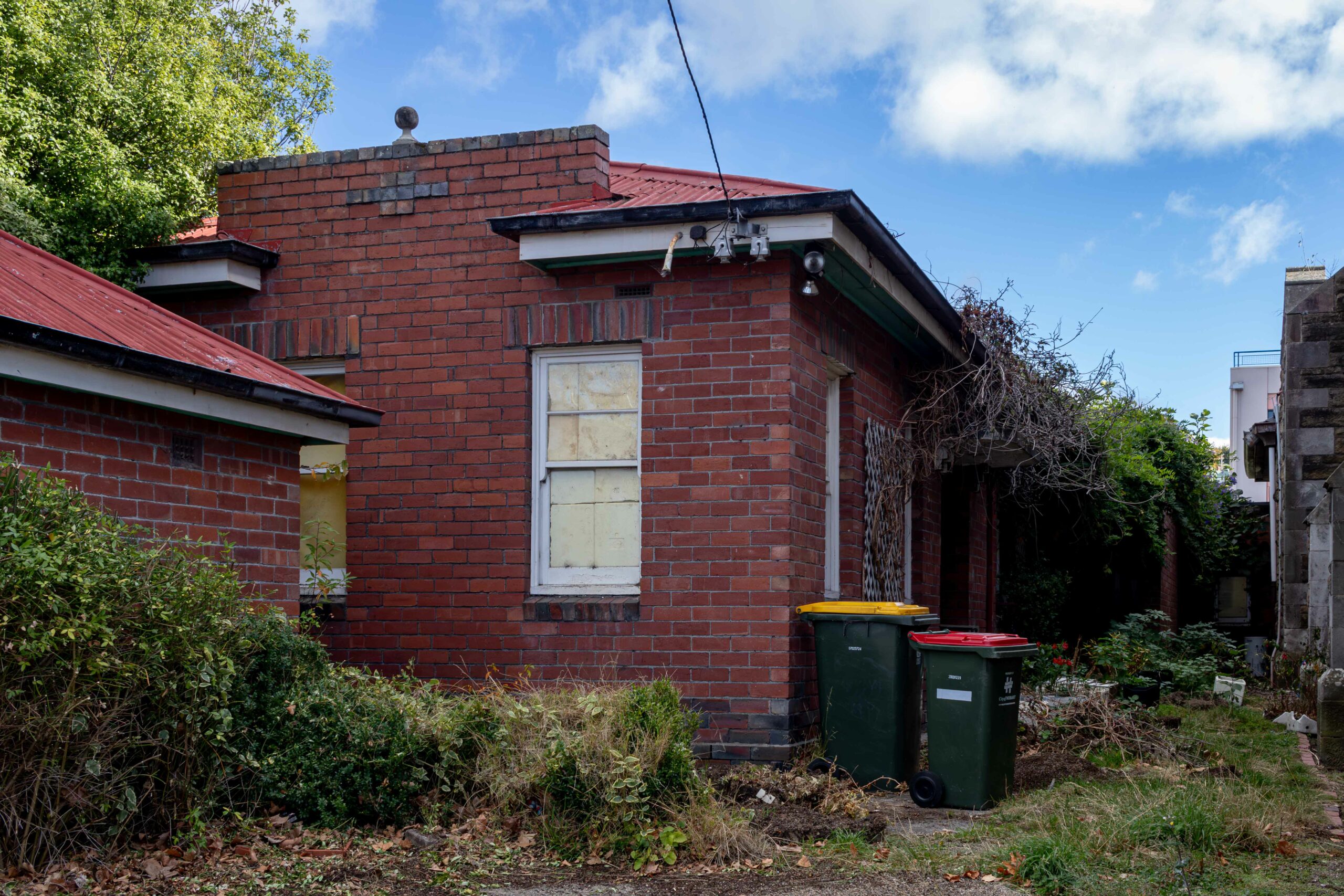 A single storey red brick house with two bins out side and a lot of green waste along the side. The windows are papered over