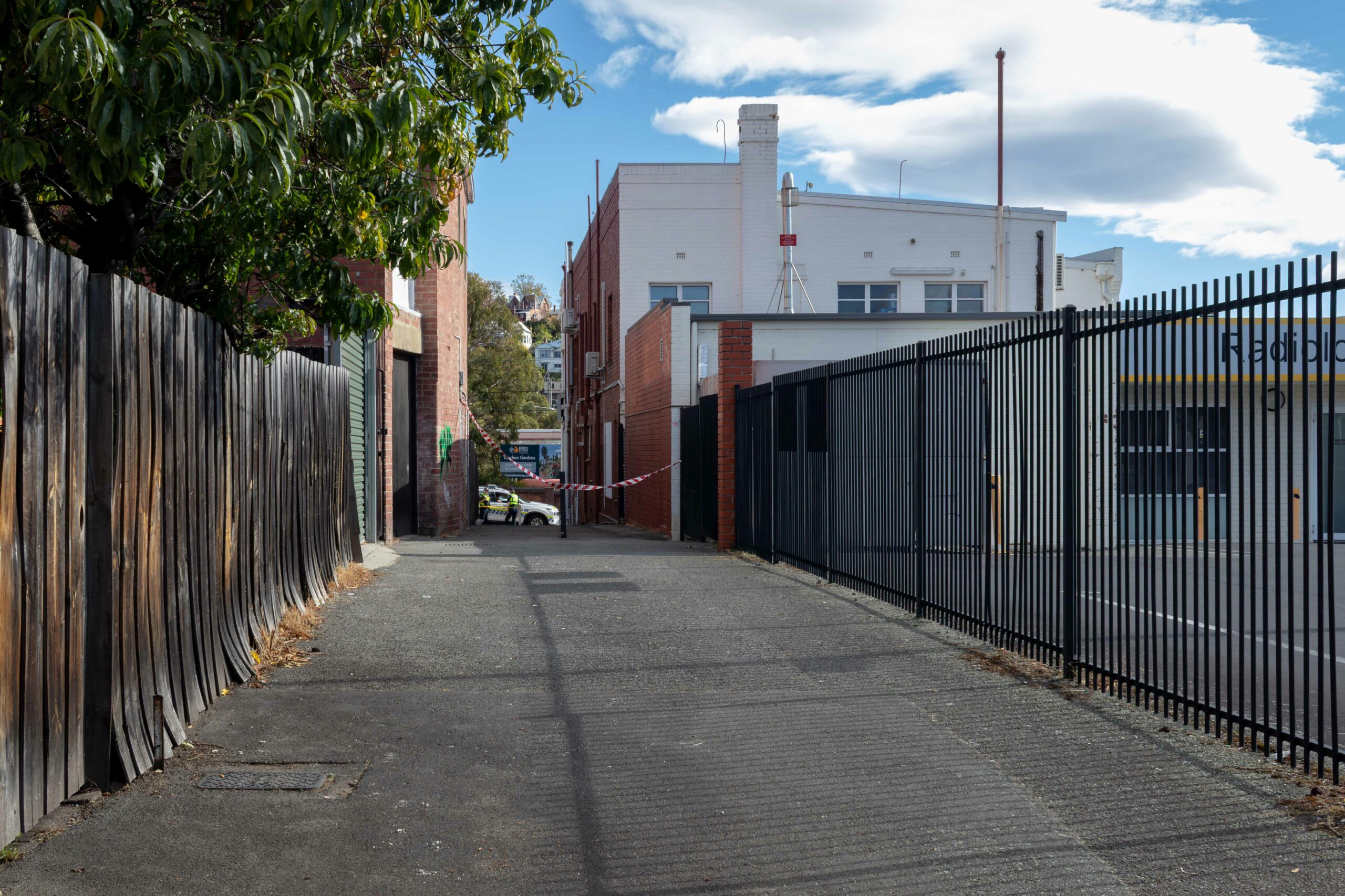 A fenced laneway with a large white bulding ot the righ tof the photo. The lane is taped off and a police car can be seen on the road at the end of the lane