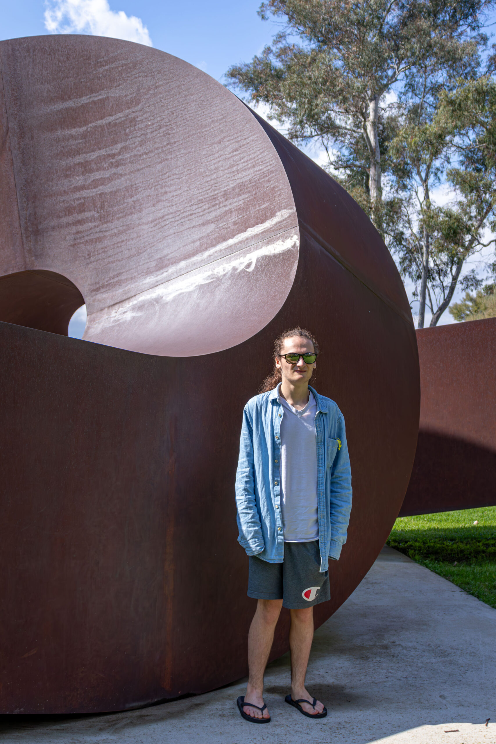 A young man in a blue shirt and sunglasses standing in front of a large round metal scultpture. He has long hair tied back