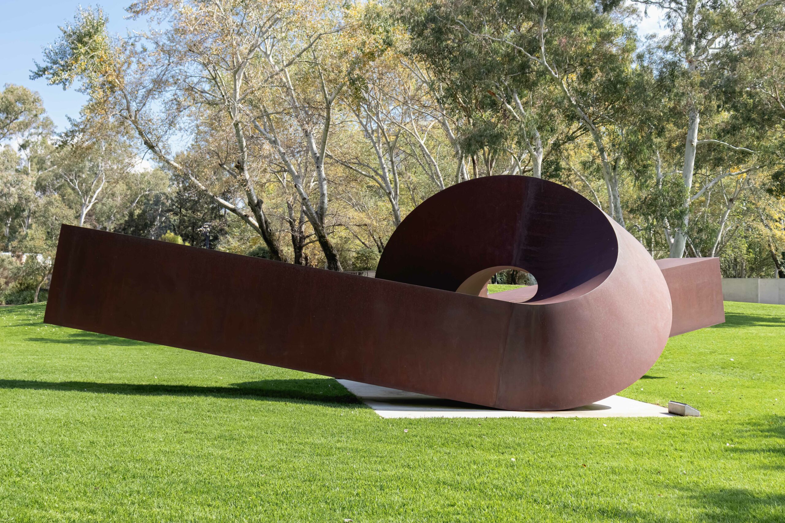A large brown metal sculpture in the shape of a twist in an outdoor setting with trees in the background